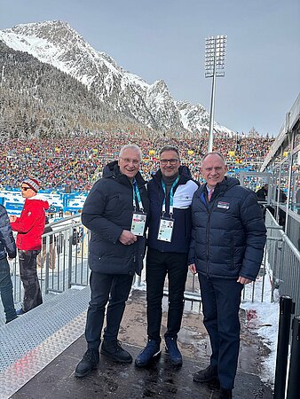 Drei Personen in dunklen Winterjacken mit Akkreditierungen stehen vor einem vollen Biathlon-Stadion und schneebedeckten Bergen.