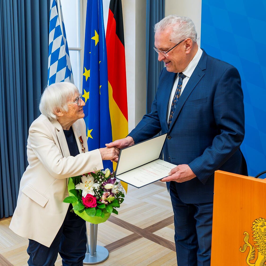 Innenminister Joachim Herrmann gratuliert händeschüttelnd Waltraud Pfandlbauer, beide stehen links von einem Rednerpult vor drei Fahnen (Bayern, Deutschland und Europa)