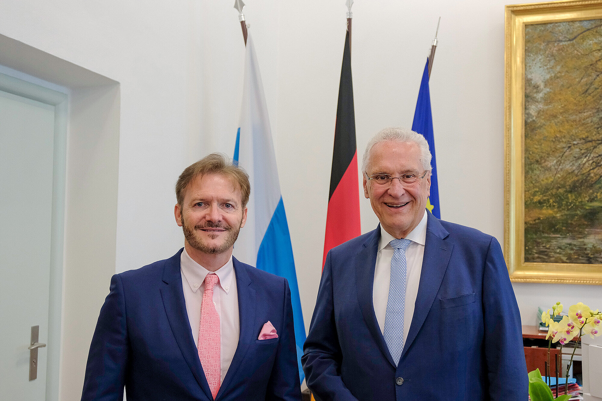 inks im Bild Generalkonsul der Schweiz Eros Robbiani, rechts im Bild Innenminister Joachim Herrmann im Ministerbüro des StMI vor einem Fahnenset