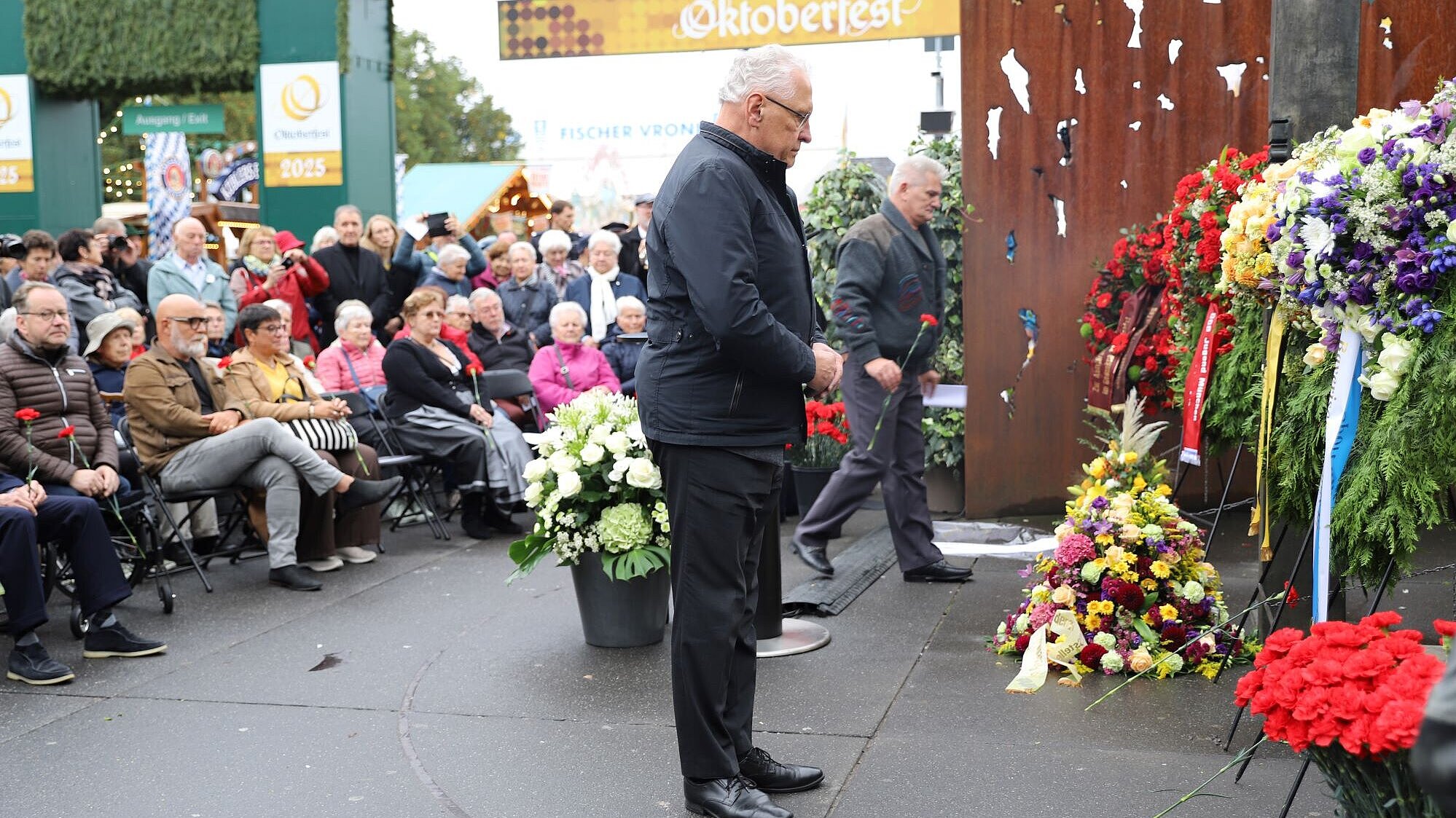 Auf dem Bild sieht man Innenminister Herrmann vor dem Gedenkkranz stehen. Links von ihm sitzen viele Leute, die auch der Gedenkveranstaltung beiwohnen.