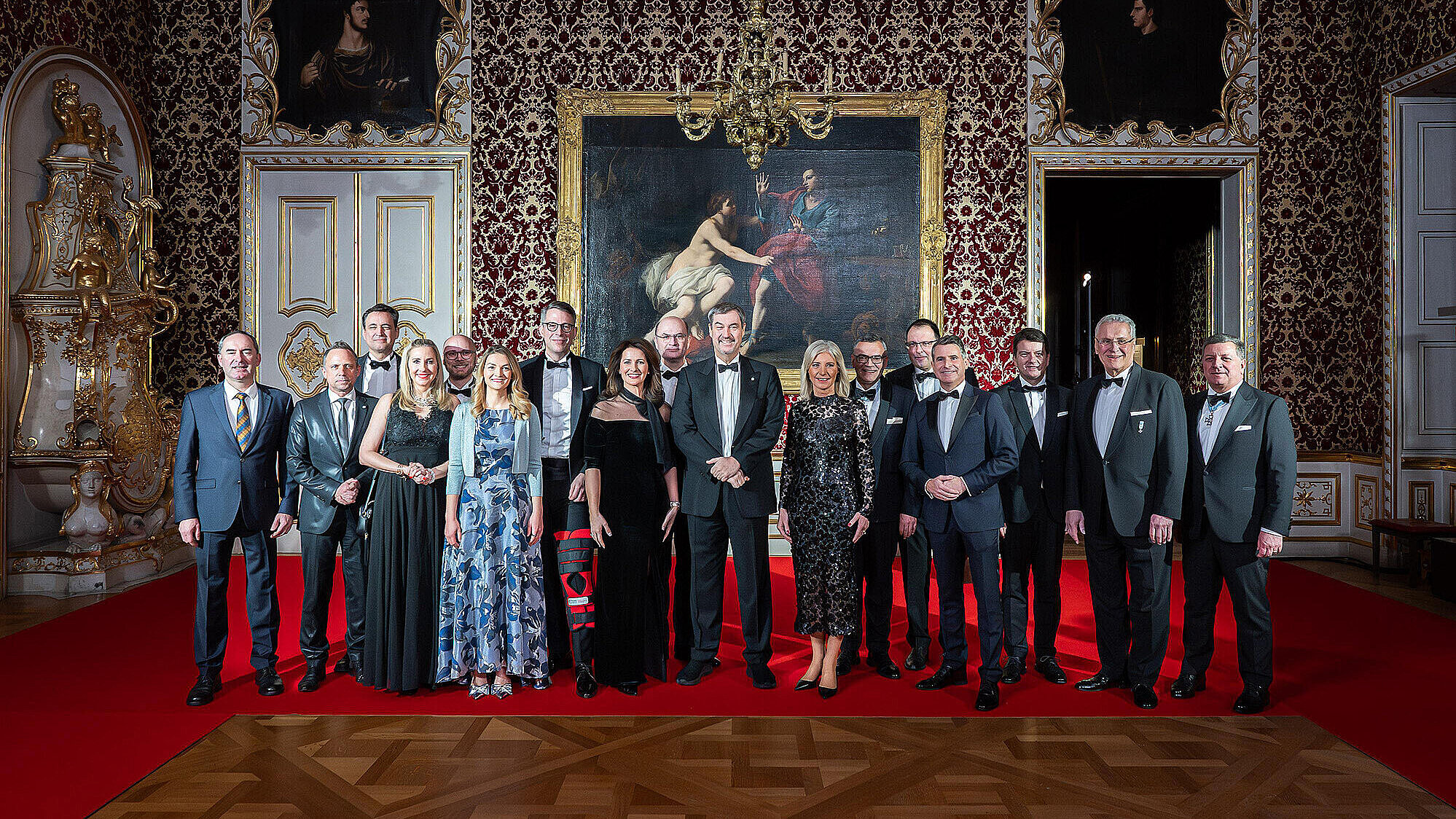 Eine Gruppe von Menschen (Mitglieder des Bayrischen Kabinetts) in festlicher Abendgarderobe steht in einem prunkvollen, historisch dekorierten Raum mit rotem Teppich und kunstvoll gemusterten Wänden. Im Hintergrund hängt ein großes, klassisches Gemälde, und eine goldene Kronleuchterleuchte ist von der Decke herab. Die Personen posieren für ein Gruppenfoto, Männer tragen Anzüge oder Smokings, Frauen elegante Kleider. Der Raum wirkt sehr repräsentativ und edel.