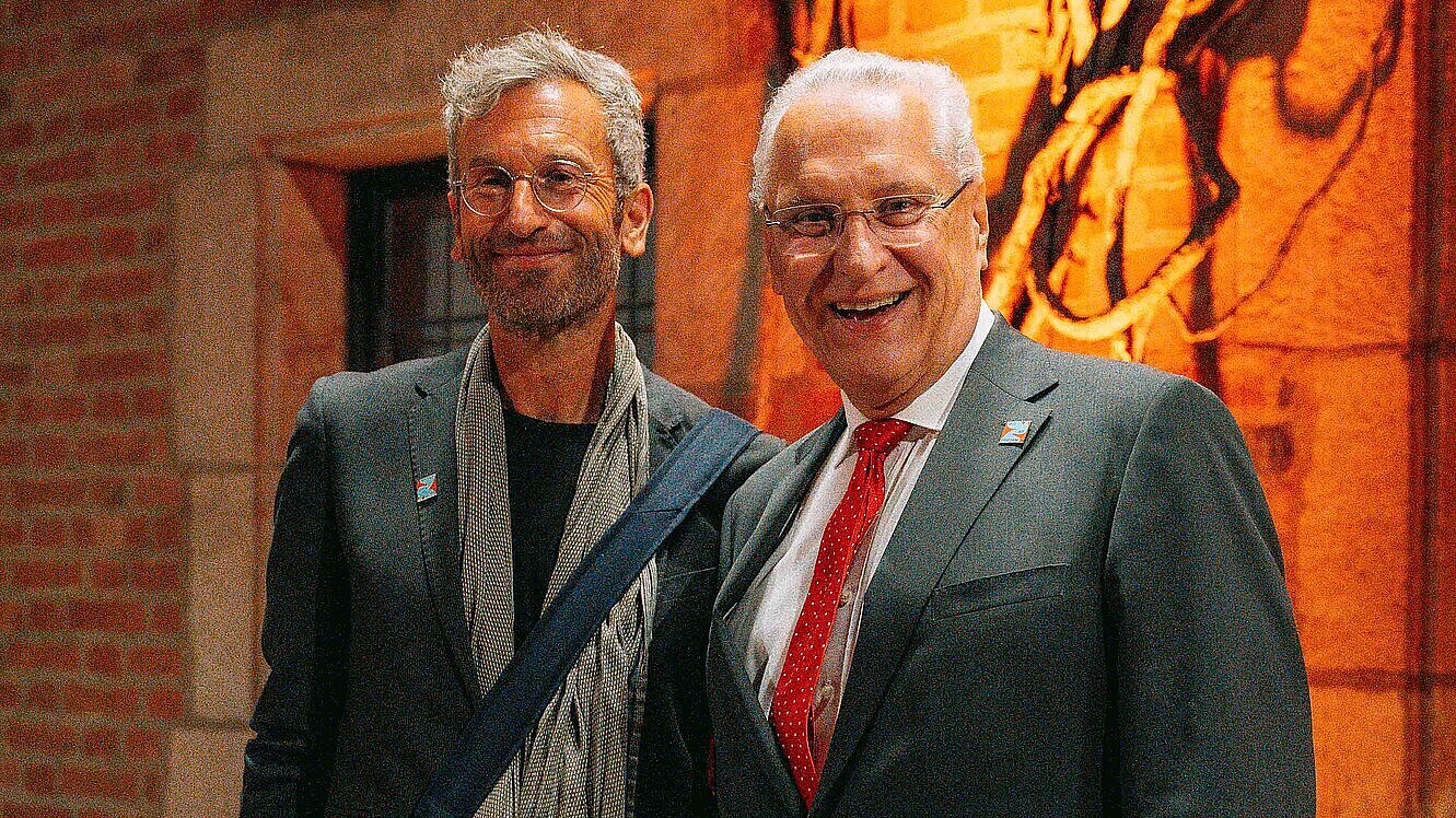 Innenminister Joachim Herrmann steht neben Moritz Puschke, dem geschäftsführenden Intendant des Musikfests ION. Vor ein beleuchteten Steinmauer.