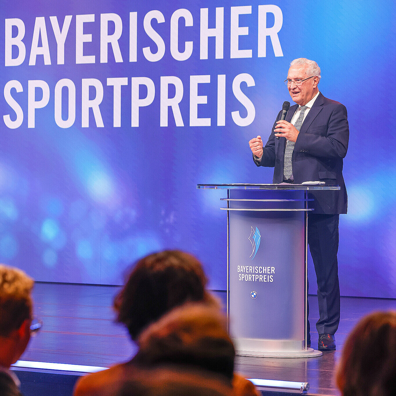Sportminister Joachim Herrmann steht am Rednerpult und spricht ins Mikrofon, im Vordergrund Publikum von hinten