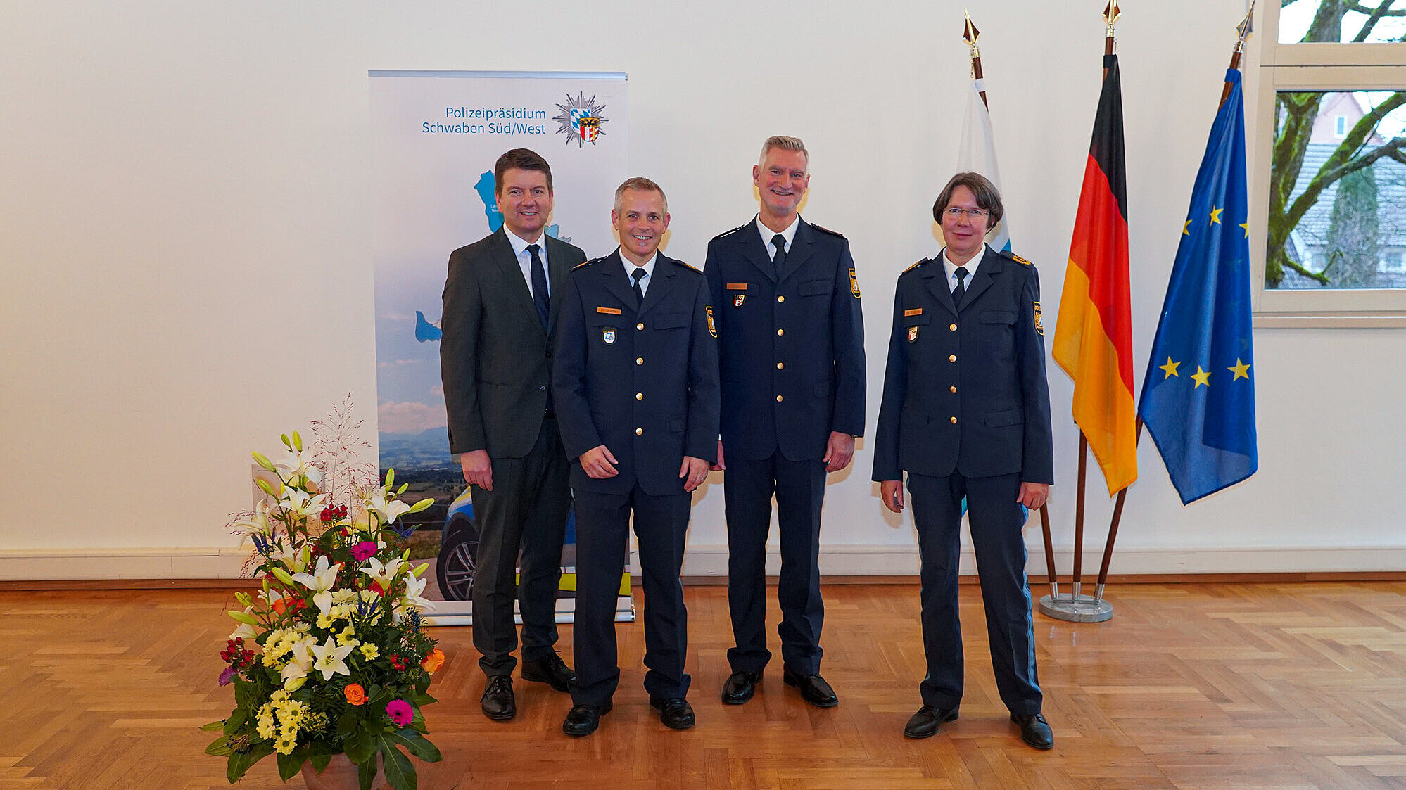 Vier Personen (von li. nach re. Innenstaatssekretär Sandro Kirchner, Dr.  Stadler, Michael Haber, Dr. Strößner)  stehen nebeneinander in einem Raum mit Parkettboden. Drei von ihnen tragen dunkelblaue Polizeiuniformen, eine Person trägt einen dunklen Anzug mit Krawatte. Im Hintergrund sind drei Fahnen zu sehen: die bayerische, die deutsche und die Europaflagge. Links steht ein großer Blumenstrauß, und hinter den Personen ist ein Banner mit der Aufschrift „Polizeipräsidium Schwaben Süd/West“ zu erkennen.