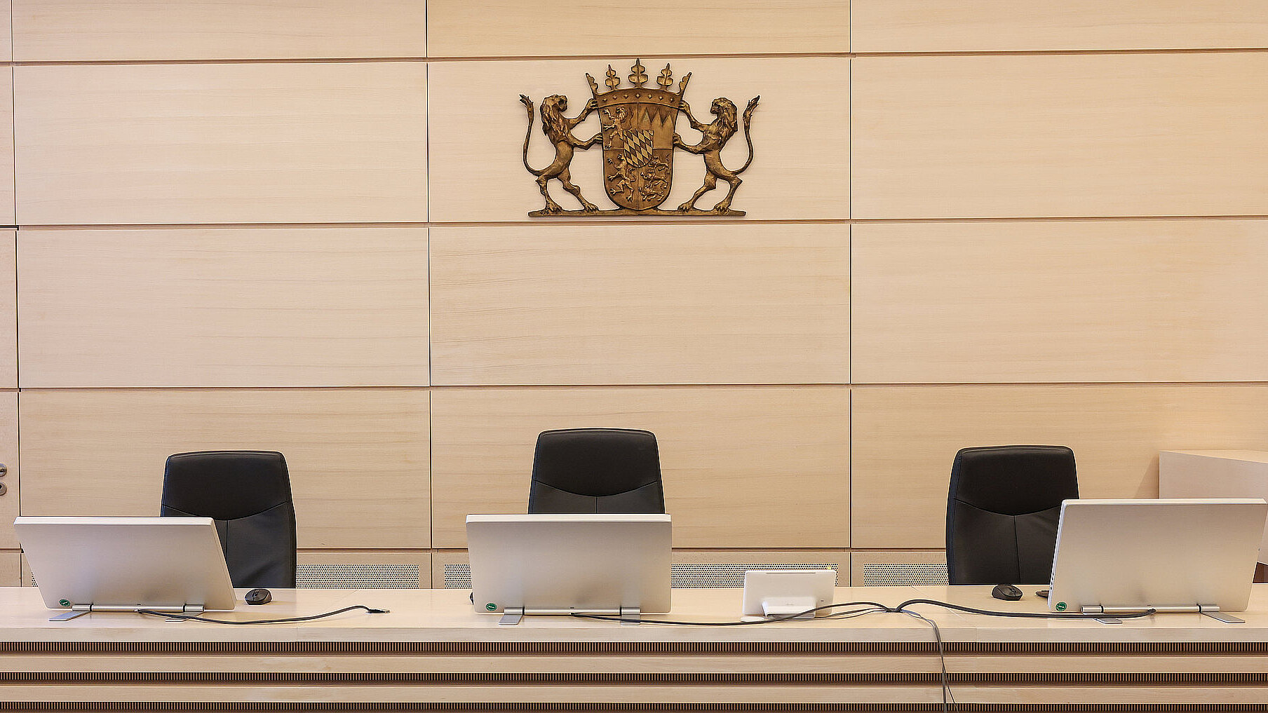 An der Wand vom Sitzungssaal des Gerichts hängt das Bayerische Staatswappen, davor sind 3 leere Sitzungsstühle