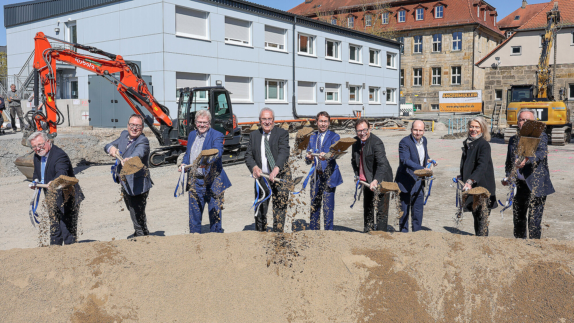 Spatenstich: mehrere Personen in Anzügen schaufeln Sand vor einer Baustelle mit Baggern und Gebäuden im Hintergrund.