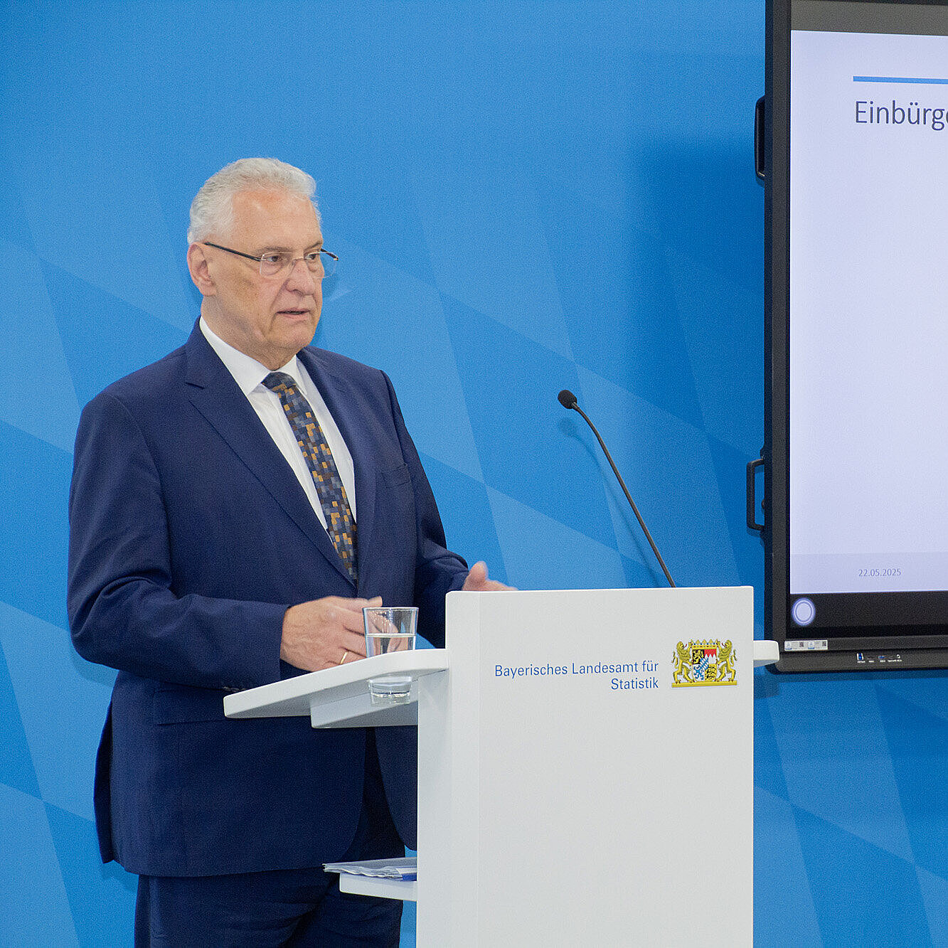 Innenminister Herrmann am Rednerpult vor blauer Pressewand im Hintergrund Bildschirm mit abgebildeter Statistik