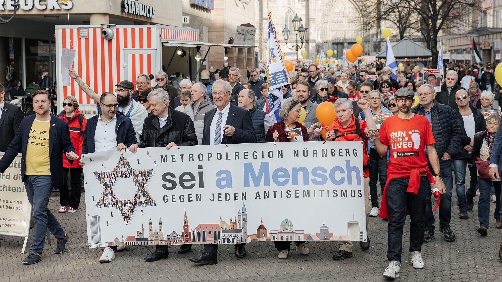 Demonstrationszug mit vielen Menschen, ganz vorne Innenminister Herrmann mit großem Schild "sei a Mensch - gegen Antisemitismus"