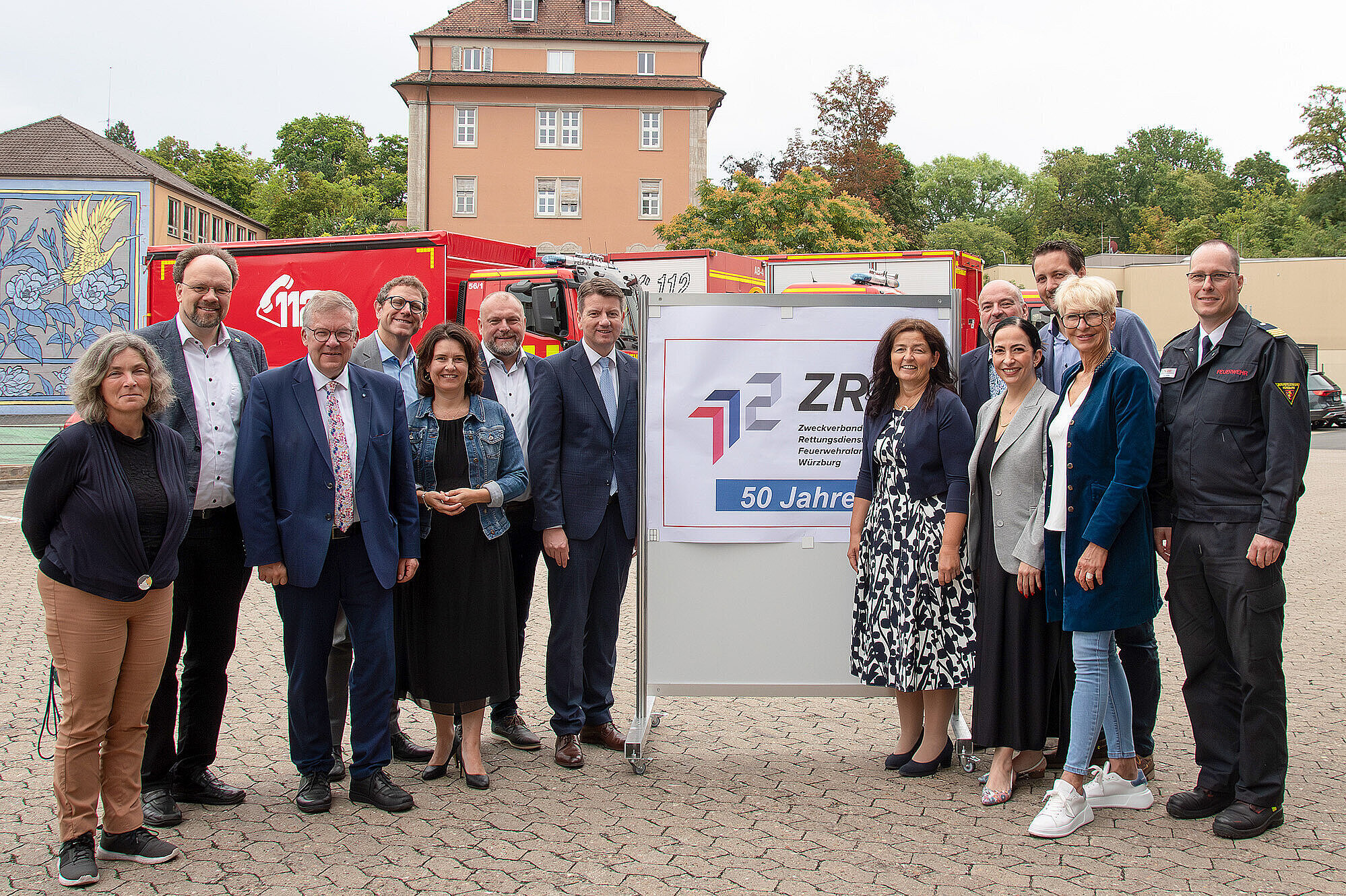 Gruppenbild von links nach rechts Ltd. Branddirektor Harald Rehmann, Landrat Thomas Eberth, Staatssekretär Sandro Kirchner, stv. Landrätin und Vorsitzende des ZRF Würzburg Christine Haupt-Kreutzer, Geschäftsführer des ZRF Würzburg Paul Justice vor einem roten Feuerwehrauto im Hintergrund ein Haus und ein Plakat auf dem ZRF steht.