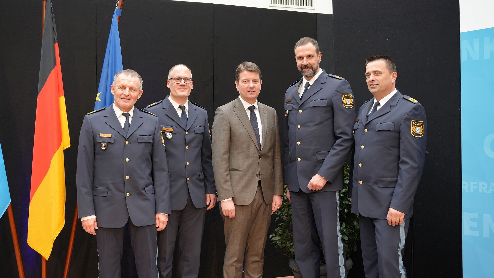 Fünf Personen auf einer Bühne, links von ihnen Fahnen. Alle fünf Personen schauen zum Fotografen.