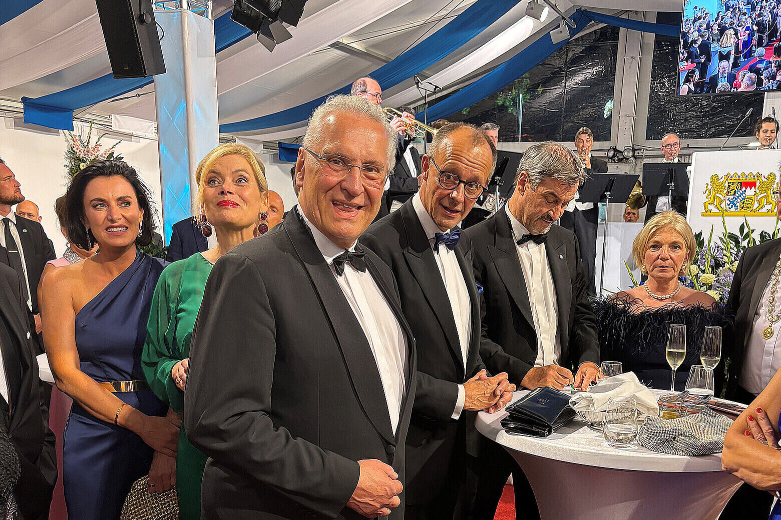 Innenminister Joachim Herrmann steht im schwarzen Smoking neben Bundeskanzler Merz im schwarzen Smoking an einem Stehtisch mit einer weißen Husse. Rundherum Gäste aus Politik und Wirtschaft in festlicher Kleidung.