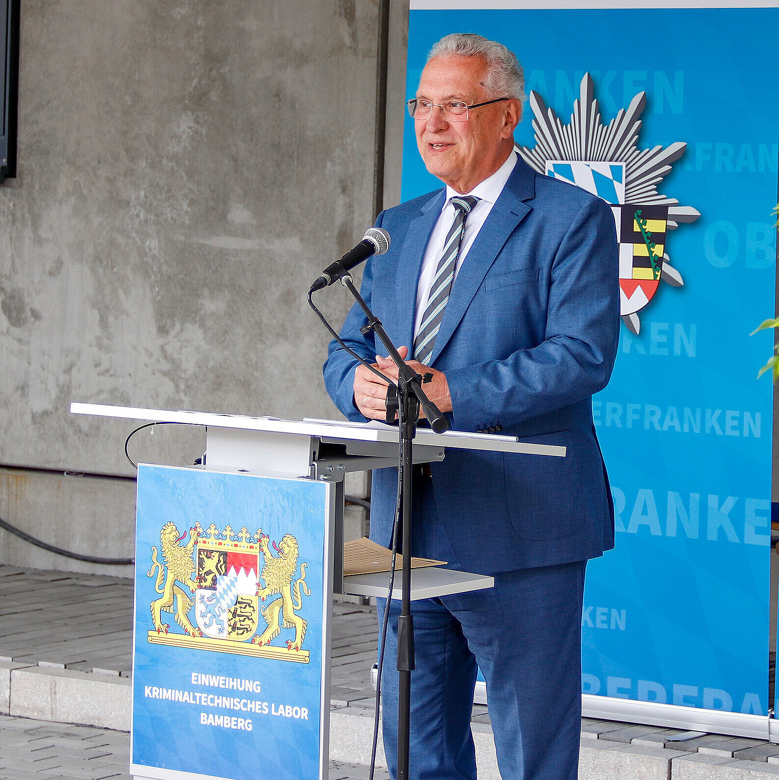Innenminister Joachim Herrmann im blauen Anzug spricht bei einer Veranstaltung am Rednerpult. Auf dem Pult ist ein Schild mit dem bayerischen Staatswappen und dem Text „Einweihung Kriminaltechnisches Labor Bamberg“ angebracht. Im Hintergrund ein Roll-Up des Polizeipräsidiums Oberfranken. Der Mann wirkt freundlich und engagiert.