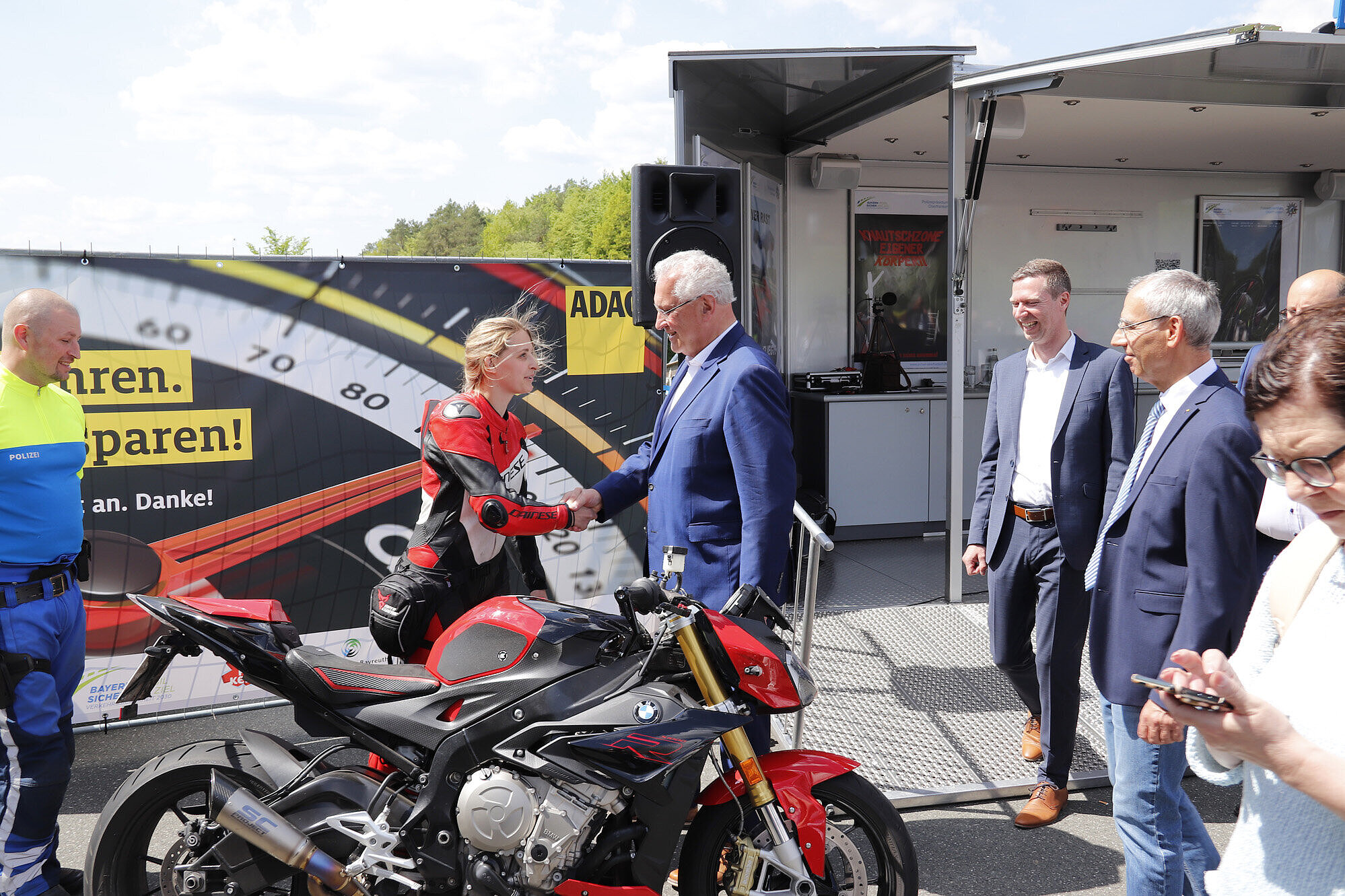 Innenminister Joachim Herrmann begrüßt eine Motorradfahrerin, im Vordergrund steht ein Motorrad.