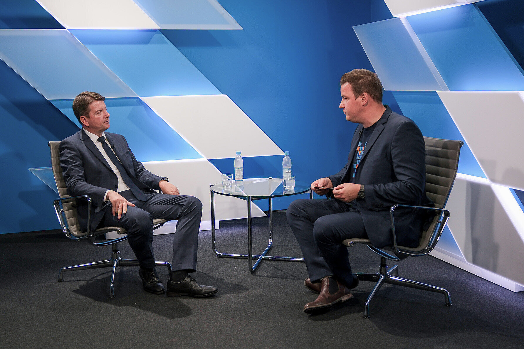 Staatssekretär Sandro Kirchner (links) im Gespräch mit einem Vertreter der BayKommun (rechts) in einem modernen Studio mit blauer, geometrischer Wandgestaltung. Beide sitzen sich in Sesseln gegenüber, ein Glastisch mit Wasserflaschen steht zwischen ihnen. 