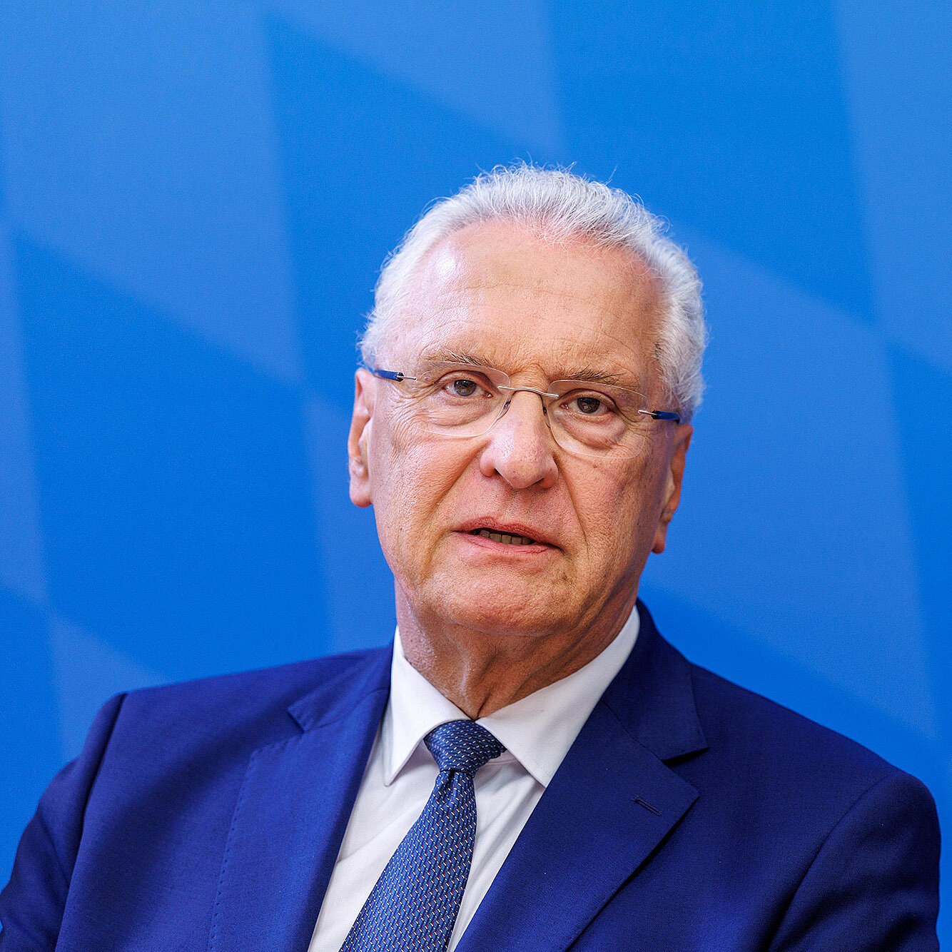 Porträt eines älteren Mannes mit weißem Haar und Brille, in dunklem Anzug und Krawatte vor einem blau gemusterten Hintergrund, ernster Gesichtsausdruck.