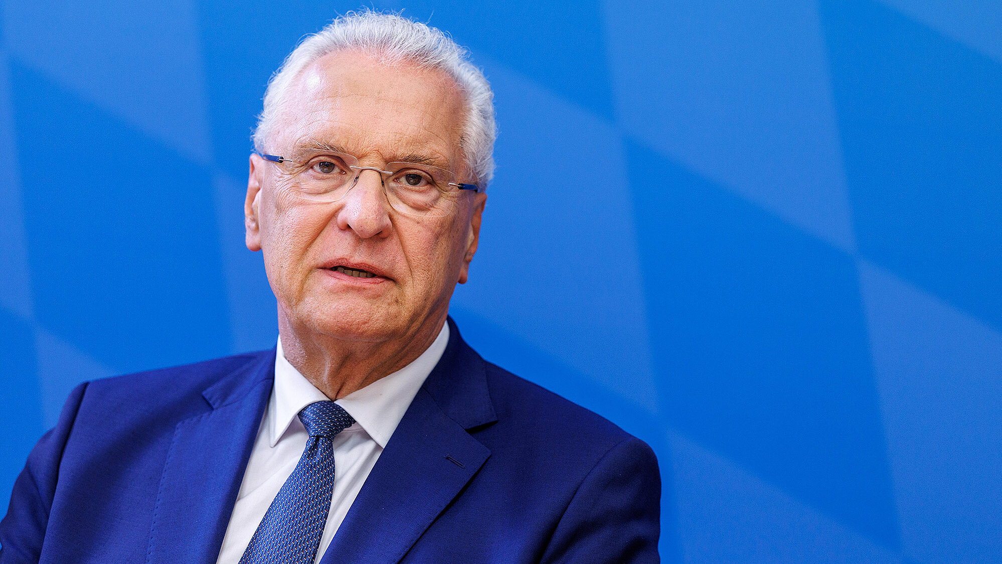 Porträt eines älteren Mannes mit weißem Haar und Brille, in dunklem Anzug und Krawatte vor einem blau gemusterten Hintergrund, ernster Gesichtsausdruck.