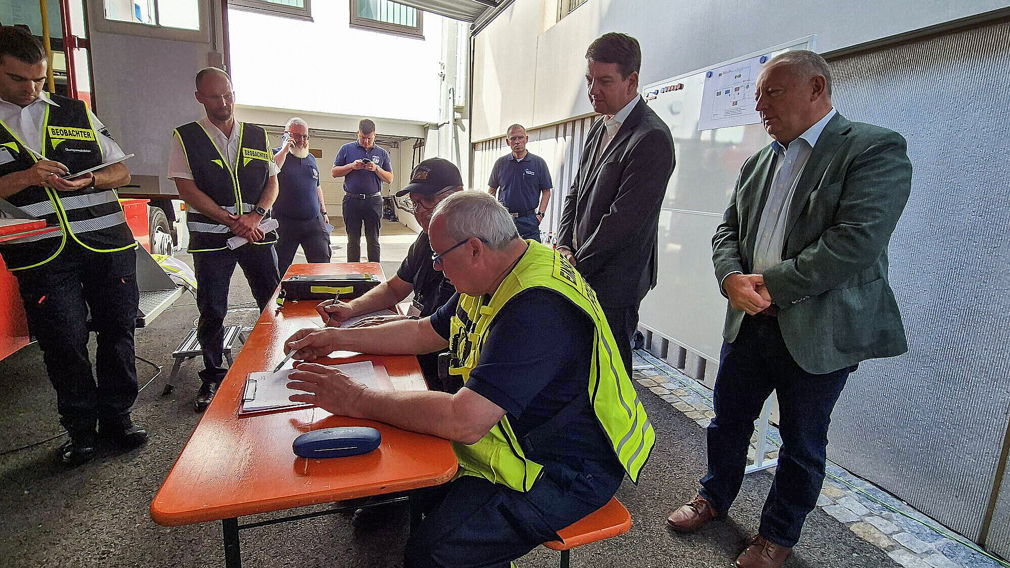 Kirchner beobachtet Menschen mit Warnweste an Bierbank mit Stift und Klemmbrett
