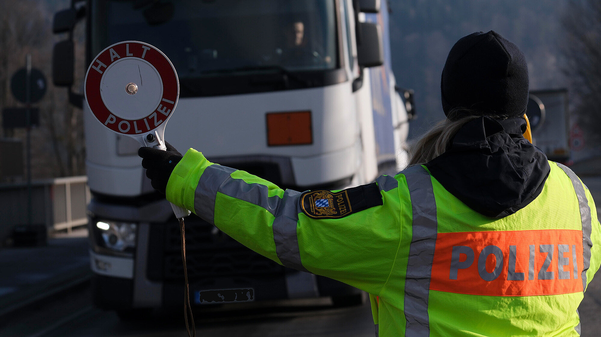 Eine Polizistin in einer leuchtend gelben Warnjacke mit der Aufschrift „POLIZEI“ auf dem Rücken hält eine Halte-Kelle mit der Aufschrift „HALT POLIZEI“ in der rechten Hand und stoppt einen weißen Lkw auf einer Straße. Der Lkw ist frontal zu sehen, die Szene spielt offenbar bei Tageslicht.