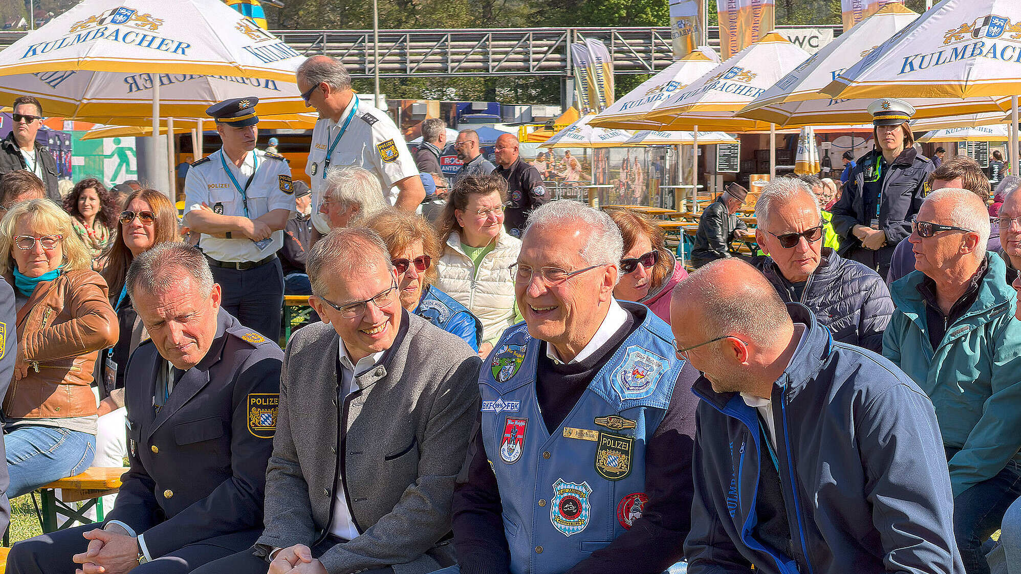 Gruppe von Menschen und Polizeibeamten sitzt auf Bierzeltbänken unter Sonnenschirmen bei einer Motorradveranstaltung im Freien.
