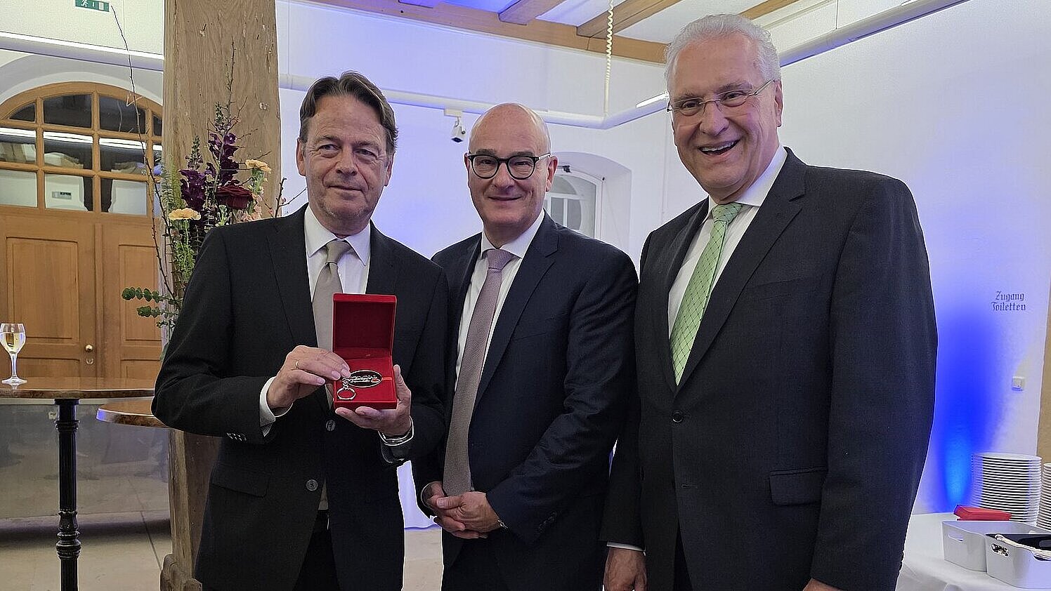 Drei Männer im Anzug nebeneinander, der linke Mann hält die Ehrenkriminalmarke in die Kamera