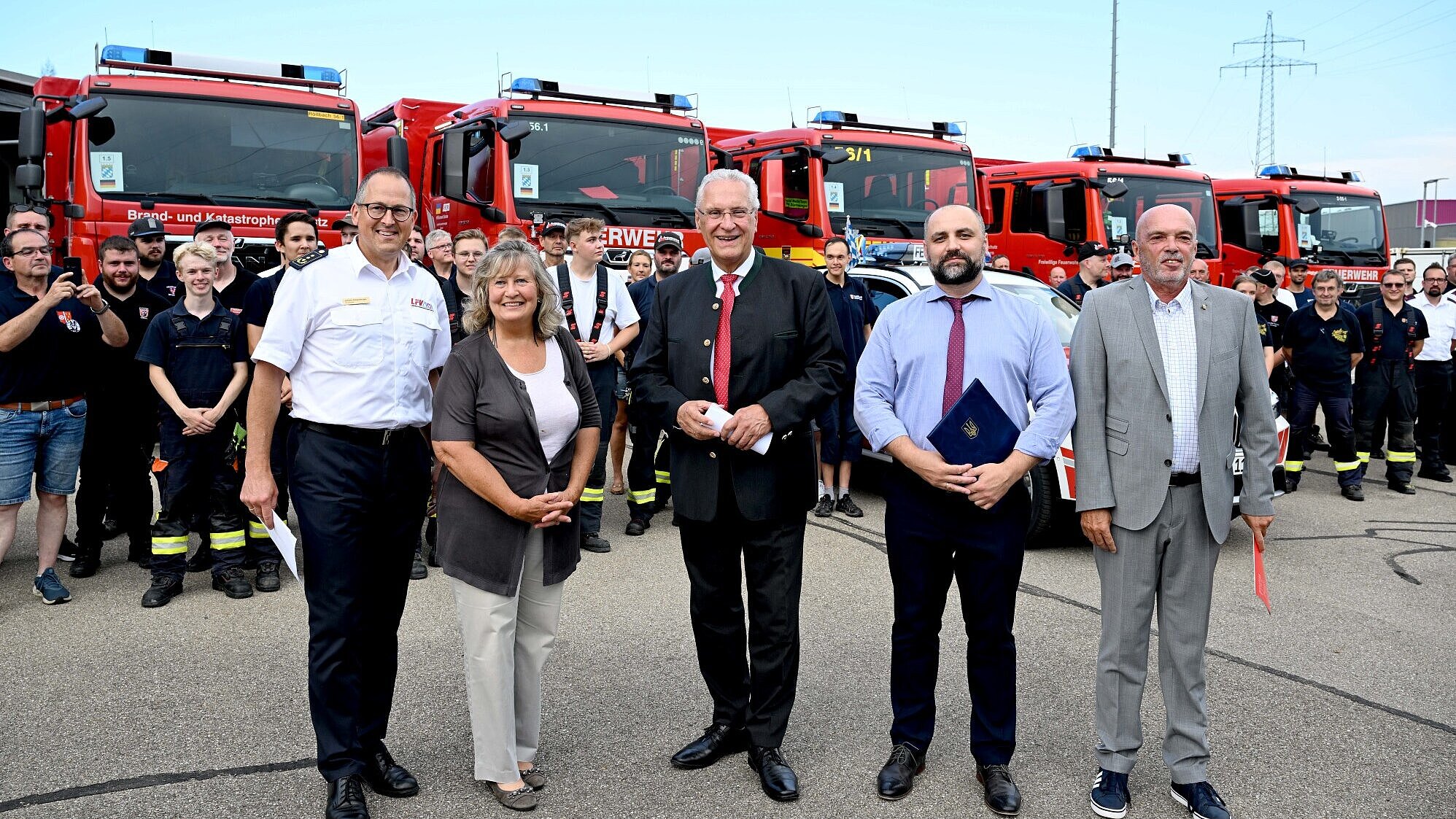 Innenminister Joachim Herrmann, ukrainischer Generalkonsul Oleksii Kravets und der Vorsitzender des Landesfeuerwehrverbandes Bayern (LFV) Johann Eitzenberger mit weiteren vor einer Gruppe Helferinnen und Helfern und Feuerwehrfahrzeuge