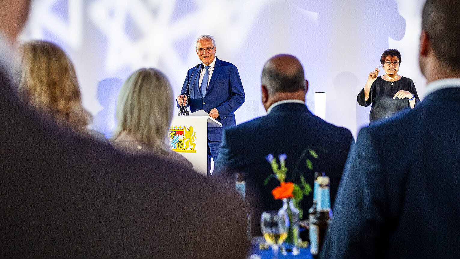 Minister Herrmann vorne auf Bühne neben Gebärdendolmetscherin, im Vordergrund Publikum an Stehtischen