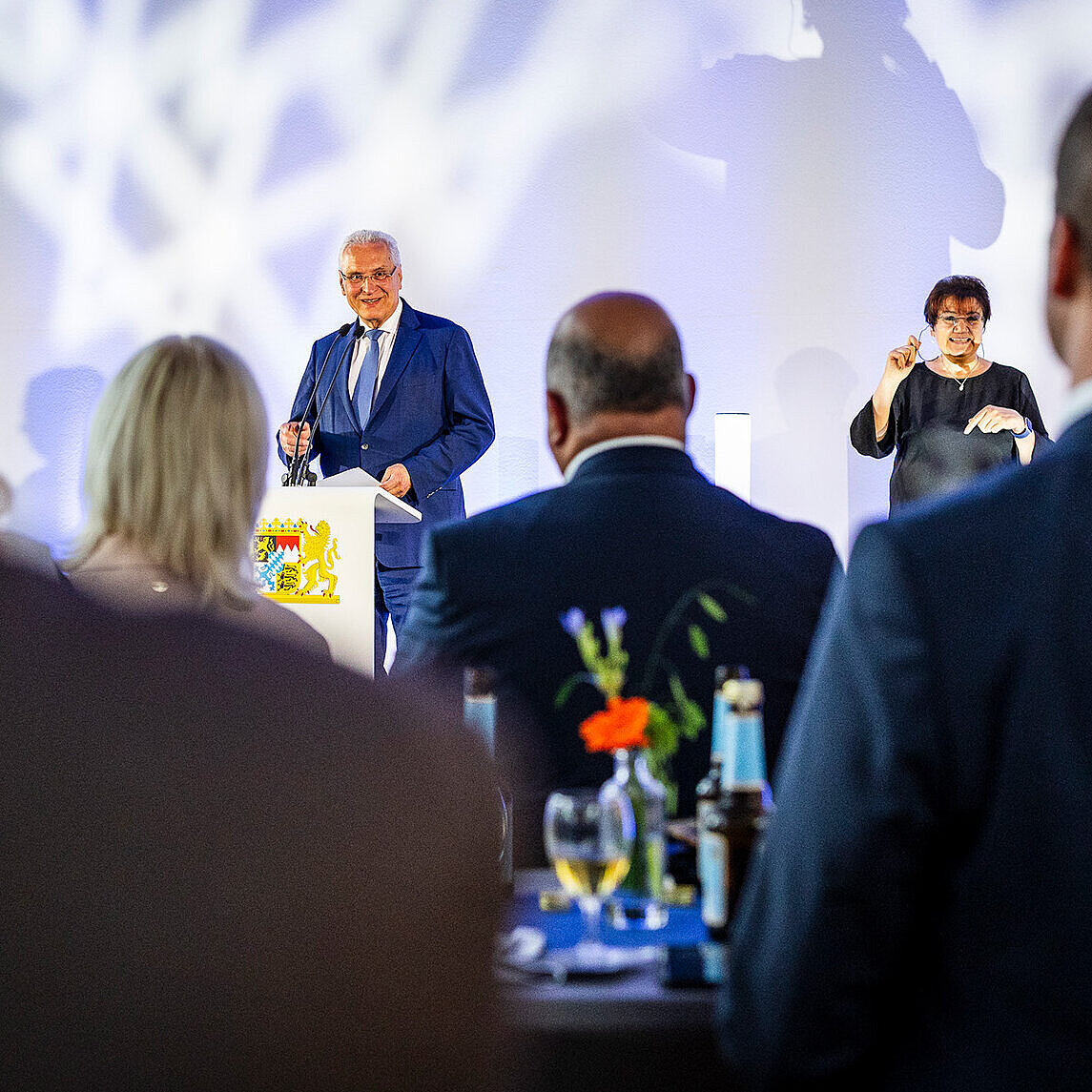 Minister Herrmann vorne auf Bühne neben Gebärdendolmetscherin, im Vordergrund Publikum an Stehtischen