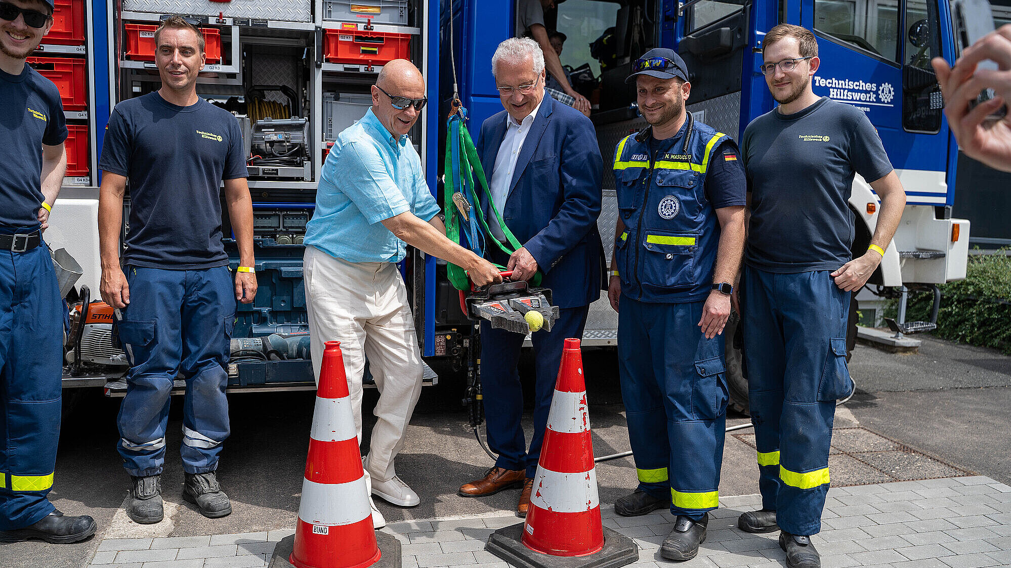 Innenminister Joachim Herrmann steht neben weiteren Herren vom THW vor einem Einsatzfahrzeug. Er absolviert einen Geschicklichkeitstest, er hält das Spreizwerkzeug des THW in der Hand und versucht einen Tennisball auf einer Pylone anzuheben.