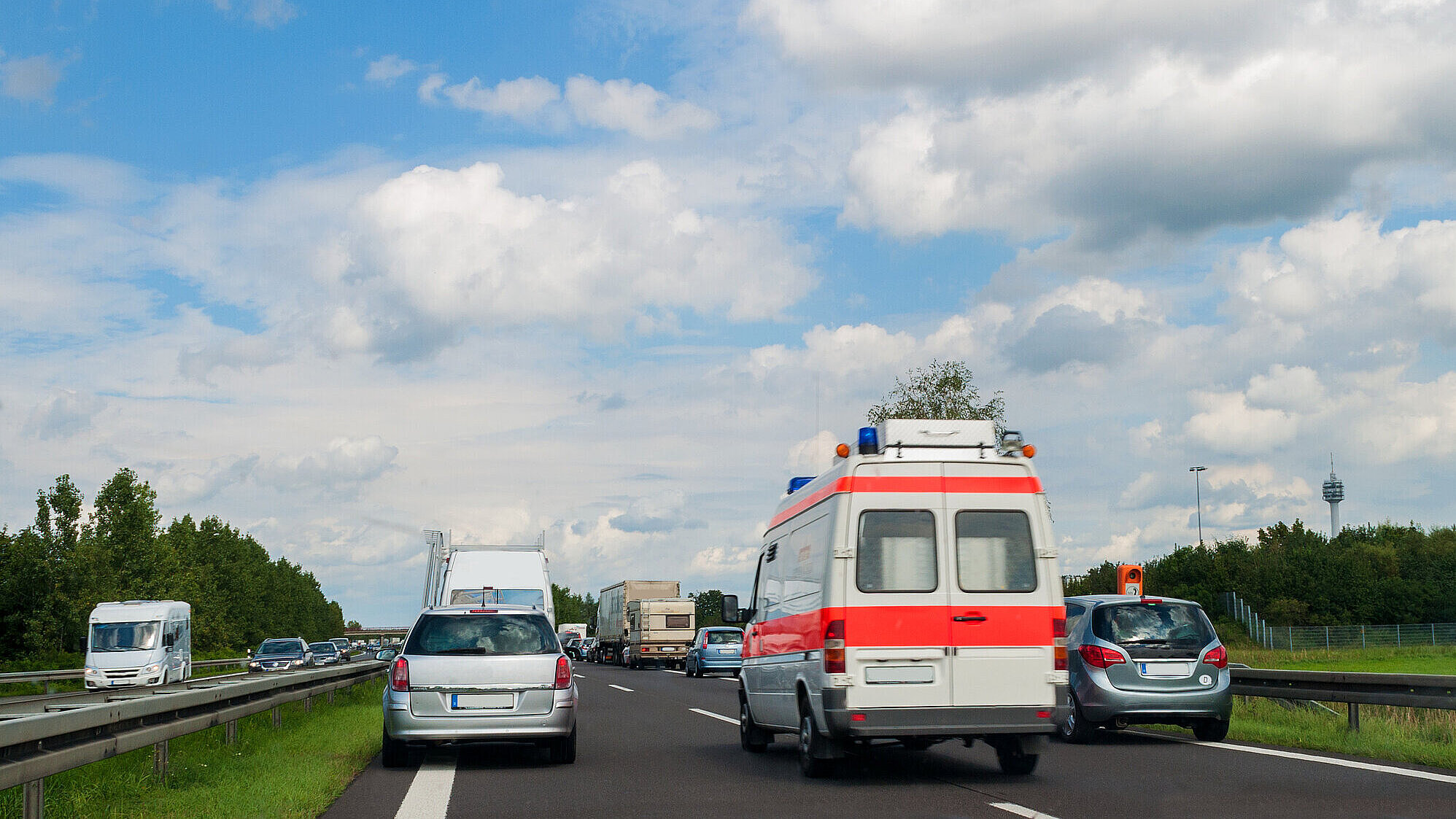 Auf dem Bild ist ein Krankenwagen zu sehen, wie dieser durch eine Rettungsgasse auf der Autobahn fährt. Links und rechts von ihm sind stehende Autos zu sehen.