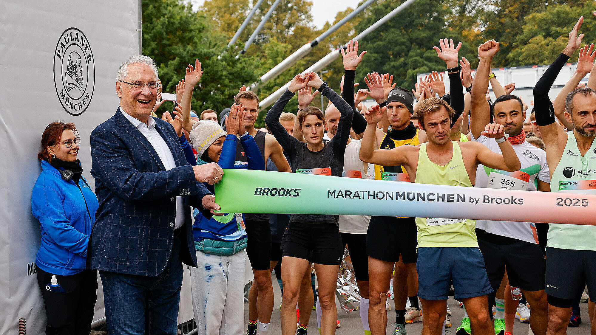 Das Bild zeigt eine Gruppe von Marathonläufern, die am Startblock des München-Marathons stehen. Ein Mann in einem blauen Anzug hält ein grünes Startband mit der Aufschrift "MARATHON MÜNCHEN by Brooks 2025" in den Händen. Die Läuferinnen und Läufer sind in sportlicher Kleidung gekleidet und scheinen sich auf den Start des Rennens vorzubereiten. Im Hintergrund sind Bäume mit herbstlicher Laubbedeckung zu sehen.