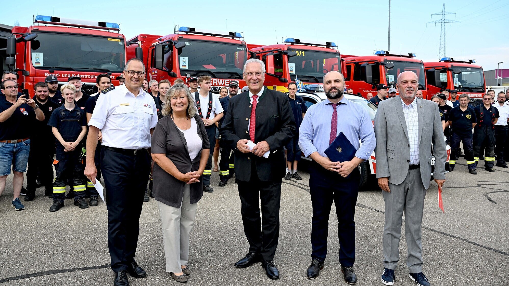 Innenminister Joachim Herrmann, ukrainischer Generalkonsul Oleksii Kravets und der Vorsitzender des Landesfeuerwehrverbandes Bayern (LFV) Johann Eitzenberger mit weiteren vor einer Gruppe Helferinnen und Helfern und Feuerwehrfahrzeuge