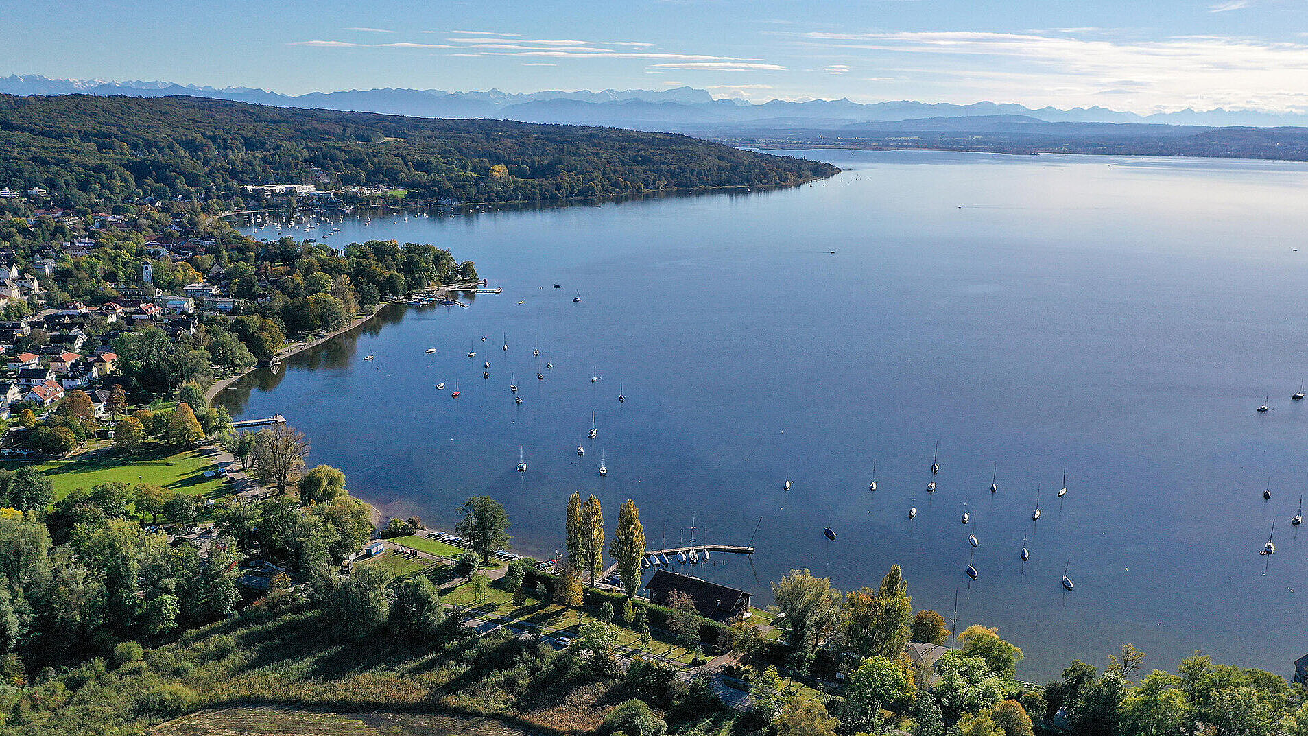Das Bild zeigt eine Luftaufnahme des Ammersees bei Herrsching. Im Vordergrund sieht man eine grüne, bewaldete Uferlandschaft mit einzelnen Gebäuden und einem kleinen Hafen mit Segelbooten. Auf dem See sind zahlreiche Segelboote verankert. Im Hintergrund erstreckt sich der See weit in die Ferne, und am Horizont sind die Umrisse einer Bergkette unter einem klaren Himmel zu erkennen.