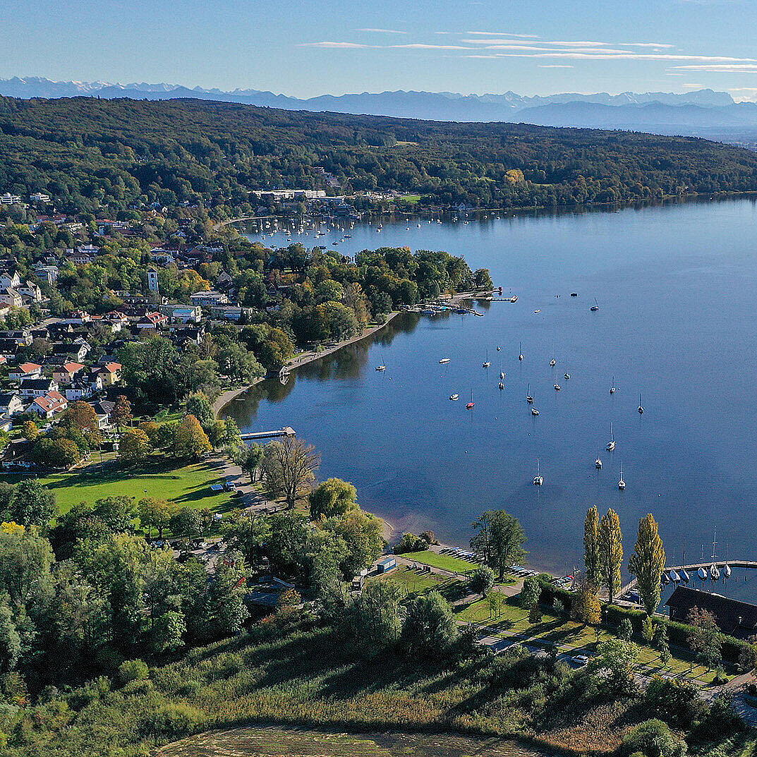 Das Bild zeigt eine Luftaufnahme des Ammersees bei Herrsching. Im Vordergrund sieht man eine grüne, bewaldete Uferlandschaft mit einzelnen Gebäuden und einem kleinen Hafen mit Segelbooten. Auf dem See sind zahlreiche Segelboote verankert. Im Hintergrund erstreckt sich der See weit in die Ferne, und am Horizont sind die Umrisse einer Bergkette unter einem klaren Himmel zu erkennen.