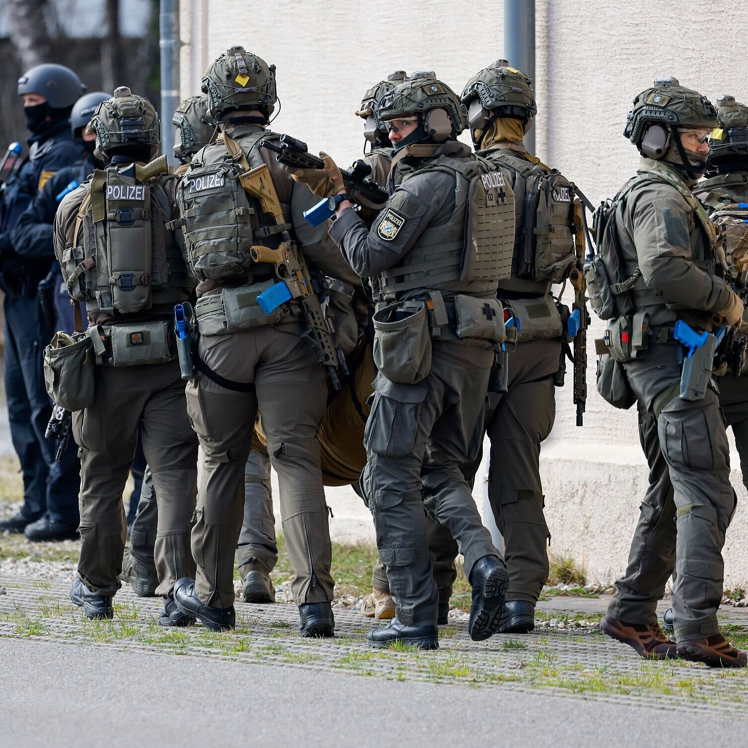 Auf dem Bild sind uniformierte Einsatzkräfte der Bayerischen Polizei und der Bundeswehr bei der Übung zu sehen. Sie stehen an einer Hausecke.