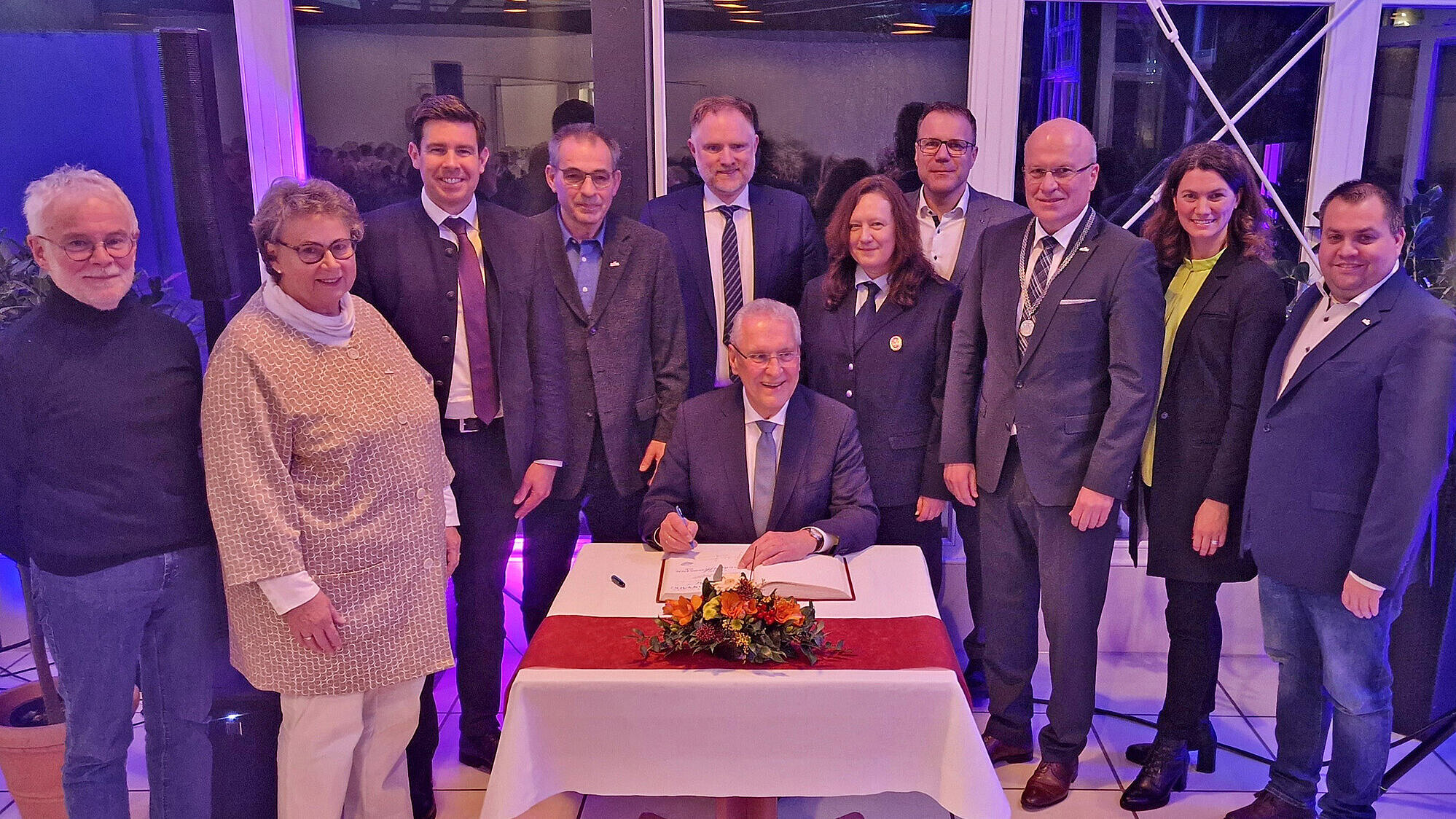 Innenminister Joachim Herrmann unterschreibt im Goldenen Buch an einem Tisch mit Blumen, umgeben von mehreren Personen in formeller Kleidung bei einem Neujahrsempfang in einem innen beleuchteten Raum.