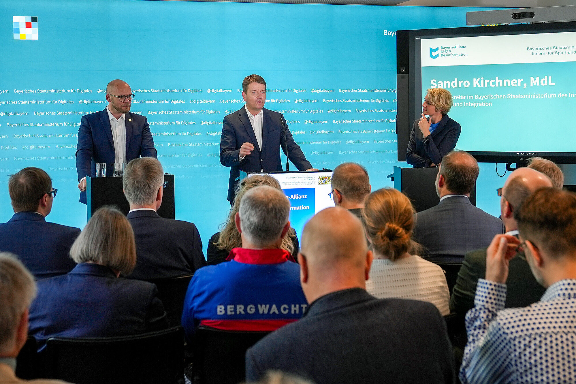 Staatssekretär Sandro Kirchner und Staatsminister Dr. Mehring vor einer blauen Pressewand mit Blick zum Publikum - rechts steht die Moderatorin