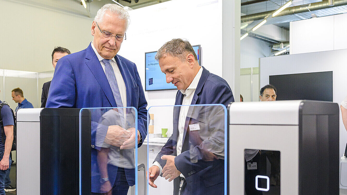 Innenminister Joachim Herrmann sieht sich eine Sicherheitsschranke mit Glastüren bei seinem Rundgang auf der SicherheitsExpo 2025 an. Ein Herr erklärt ihm die Funktionen