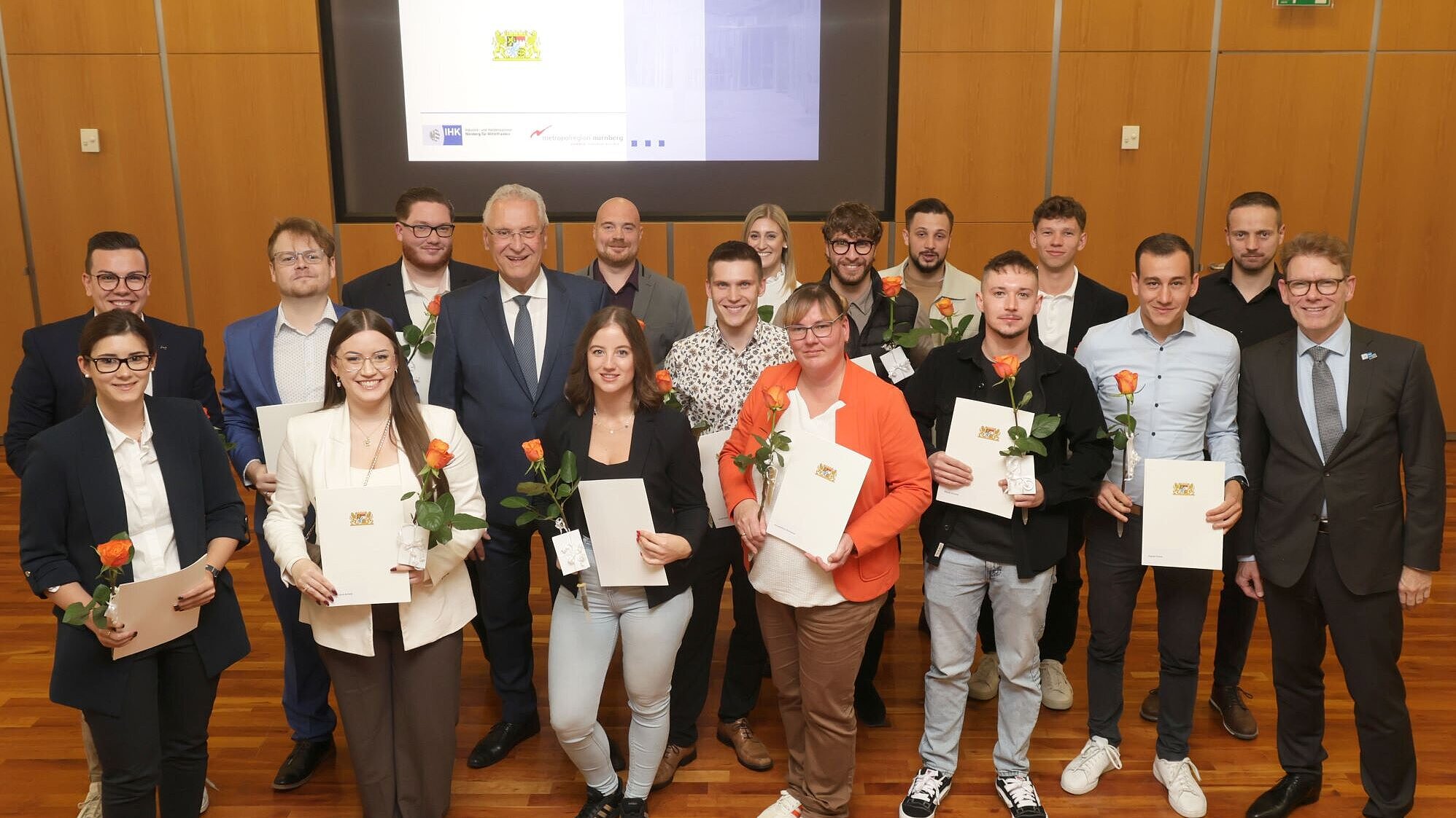 Auf dem Bild ist Innenminister Joachim Herrmann mit den Geehrten anlässlich der Meisterpreisverleihung zu sehen. Sie stehen auf einer Bühne, hinter ihnen eine Präsentation.