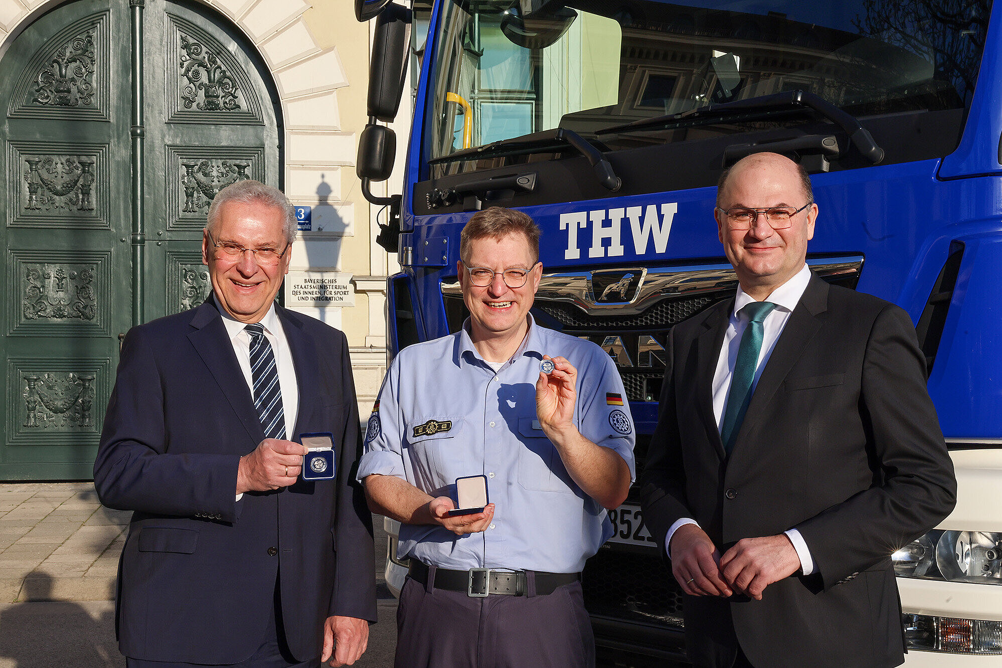 Auf dem Bild: von links nach rechts: Innenminister Joachim Herrmann, Dr. Fritz-Helge Voß, Finanzminister Albert Füracker halten die Gedenkmünze in die Kamera im Hintergrund ein THW Fahrzeug und der Eingang zum StMI