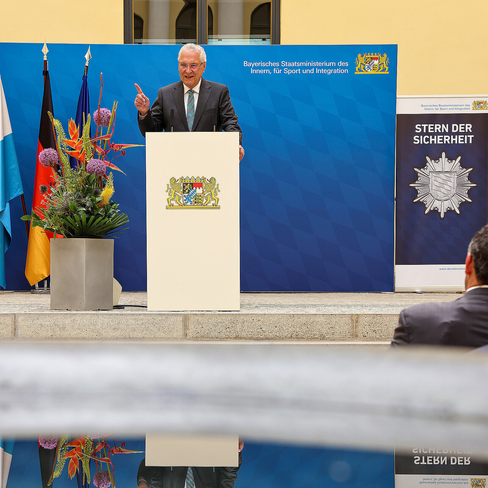 Innenminister Herrmann am Rednerpult hinter ihm steht eine blaue Pressewand links davon ein Fahnenset und rechts ein Rollup mit der Aufschrift Stern der Sicherheit