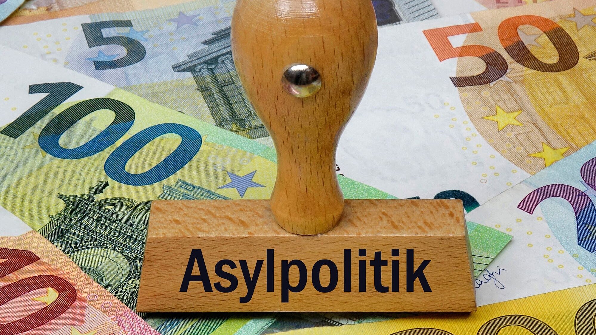 Auf dem Bild ist ein Stempel mit der Aufschrift "Asylpolitik" zu sehen, darunter liegen mehrere Geldscheine.