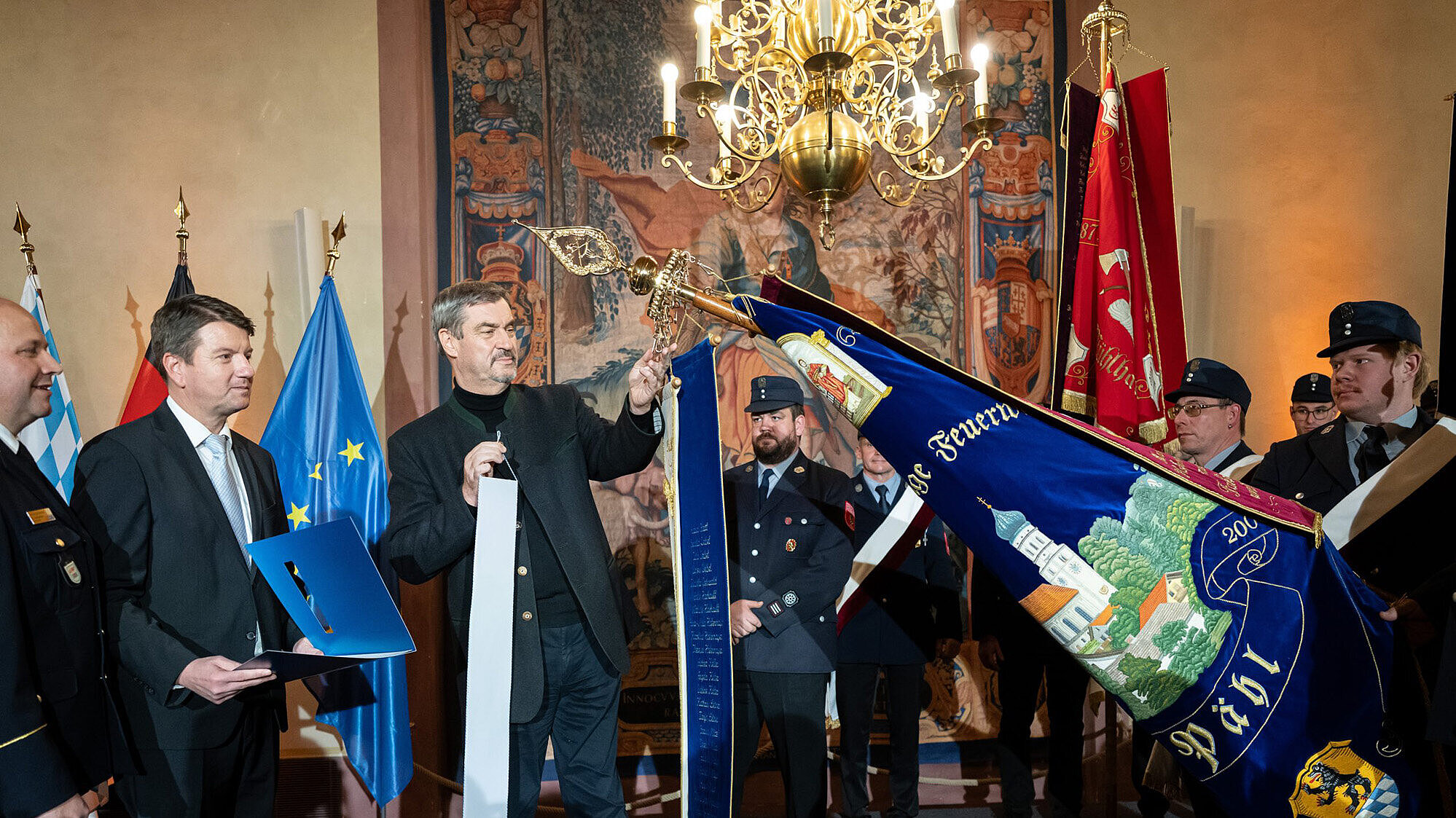 Eine feierliche Zeremonie in einem prunkvollen Raum mit Wandteppichen und einem Kronleuchter. Mehrere Personen in Uniform und Anzug stehen zusammen, eine Person hält eine große, reich verzierte Fahne mit der Aufschrift „Feuerwehr Peißenberg“. Im Hintergrund sind die Flaggen von Bayern, Deutschland und der Europäischen Union zu sehen.