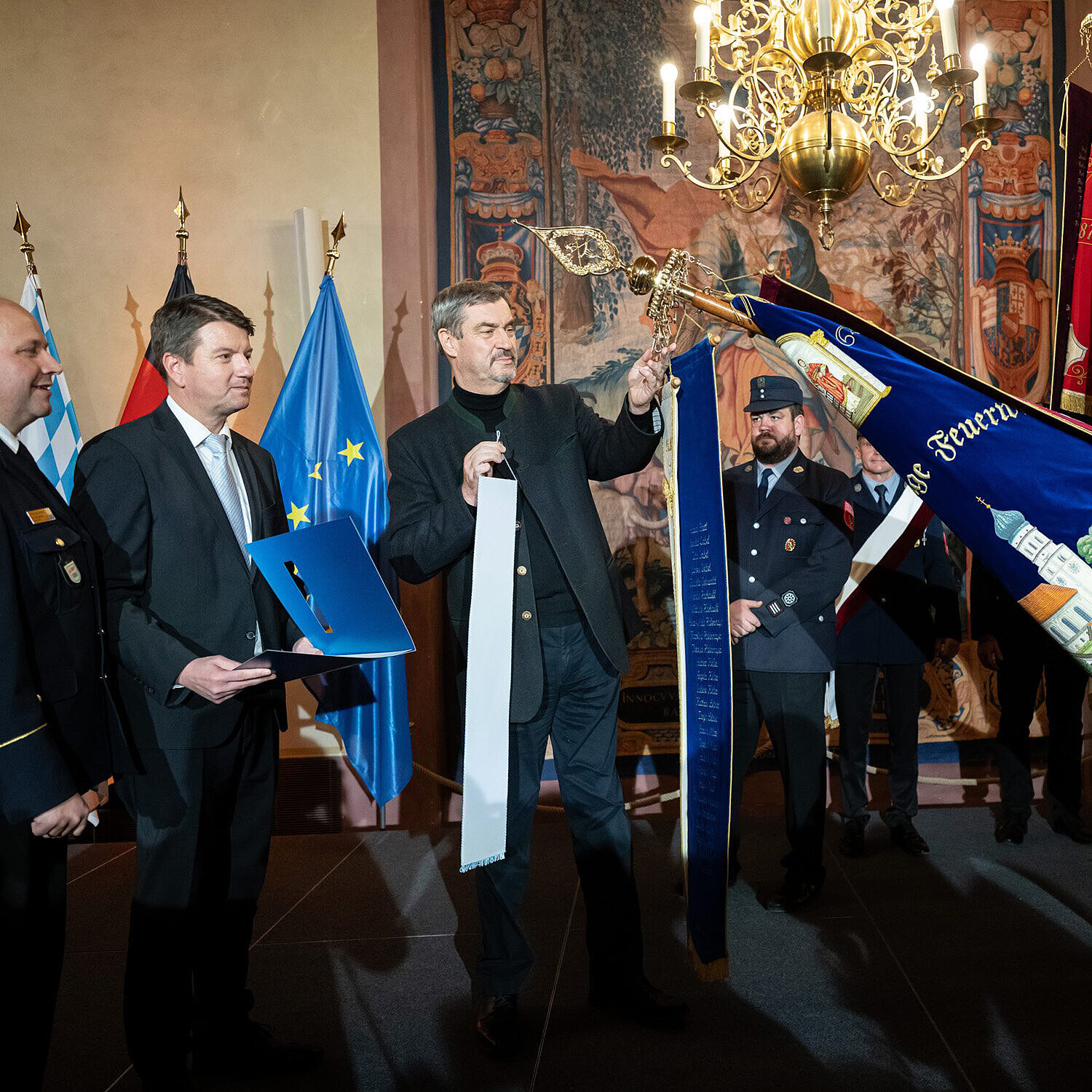 Eine feierliche Zeremonie in einem prunkvollen Raum mit Wandteppichen und einem Kronleuchter. Mehrere Personen in Uniform und Anzug stehen zusammen, eine Person hält eine große, reich verzierte Fahne mit der Aufschrift „Feuerwehr Peißenberg“. Im Hintergrund sind die Flaggen von Bayern, Deutschland und der Europäischen Union zu sehen.
