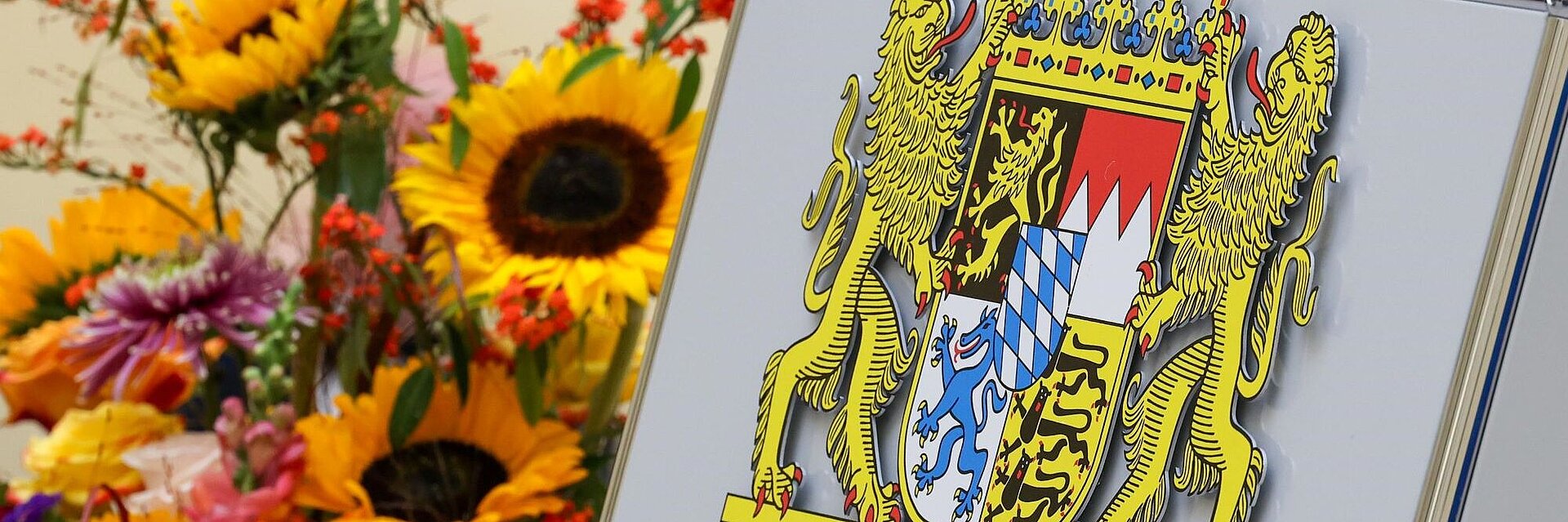 Bayerisches Staatswappen aus Metall neben Blumen