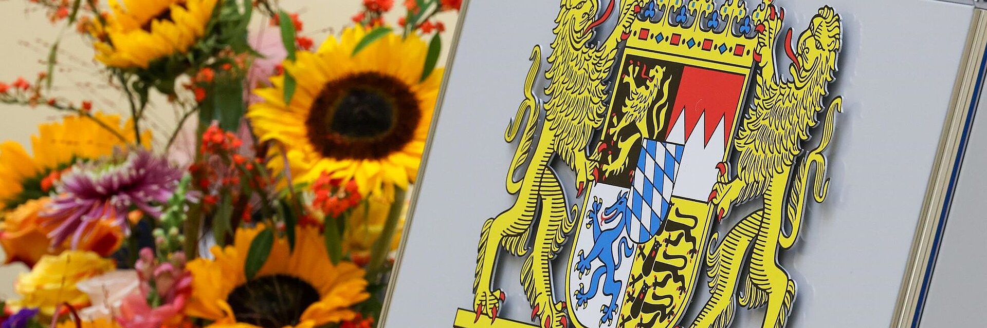 Bayerisches Staatswappen aus Metall neben Blumen