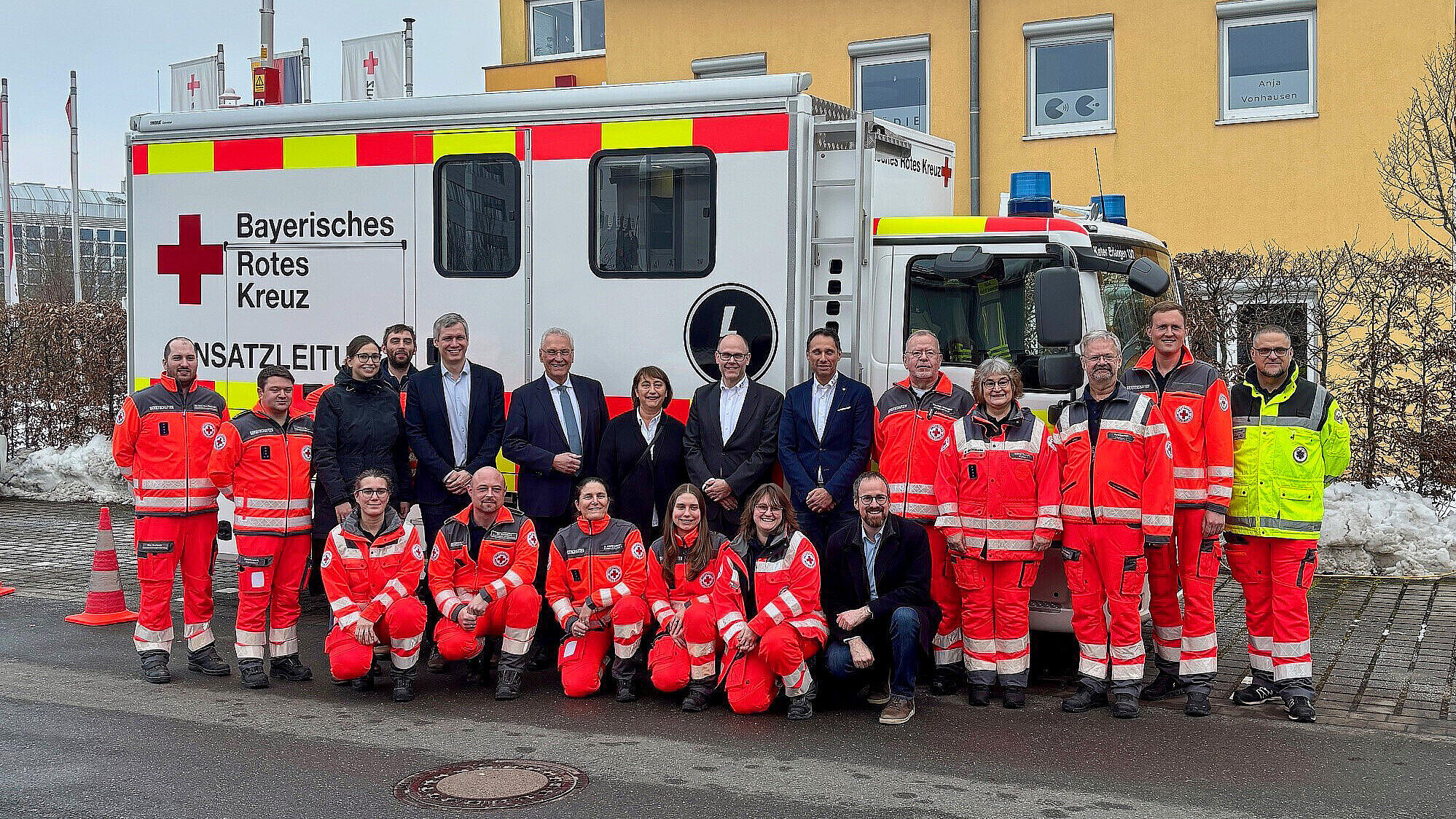 Etwa 18 Personen stehen und hocken in zwei Reihen; sie tragen überwiegend rote Rettungsdienst- bzw. Einsatzkleidung, einige wenige tragen dunkle Anzüge oder eine gelbe Warnjacke.