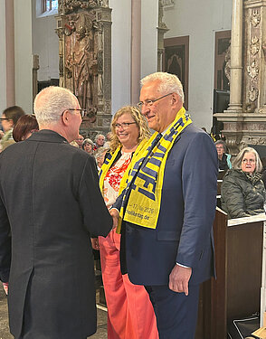 Drei Personen stehen in einer Kirche; links ein Mann mit grauem Haar von hinten, rechts ein Mann im Anzug mit gelb-blauem Schal, die sich die Hand geben; in der Mitte eine Frau mit floraler Bluse und pinker Hose. Im Hintergrund Kirchenraum mit Sitzenden. Gesichter unkenntlich.