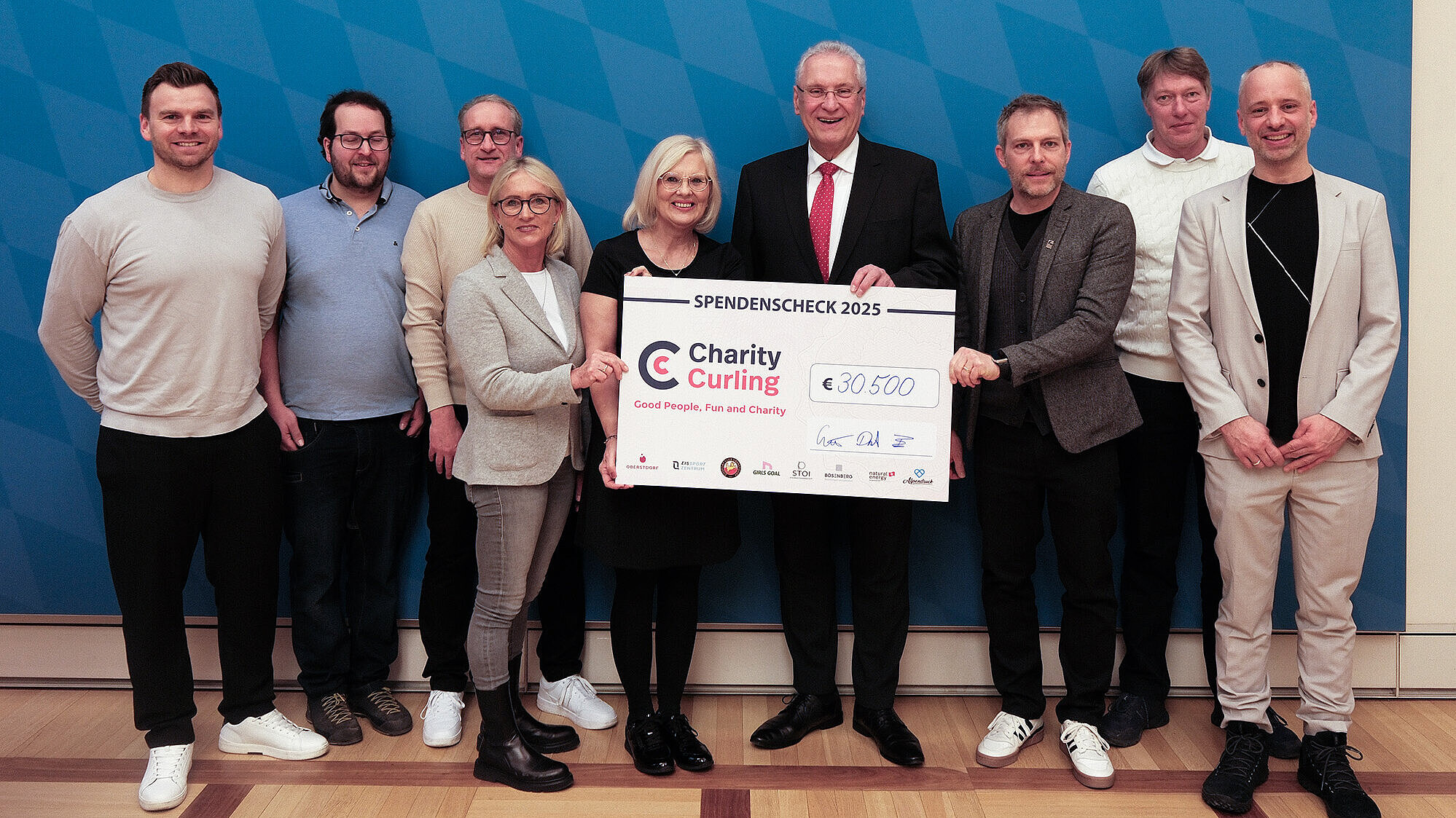Mehrere Erwachsene (4. v. li. Innenminister Joachim Herrmann) stehen eng nebeneinander vor einer Wand mit blau‑weißem Rautenmuster und halten gemeinsam einen großen Spenden‑Scheck mit dem Logo „Charity Curling“ und dem eingetragenen Betrag „€30.500“; die Personen tragen teils legere, teils formelle Kleidung, die Gesichter sind unkenntlich gemacht; Holzboden im Vordergrund.