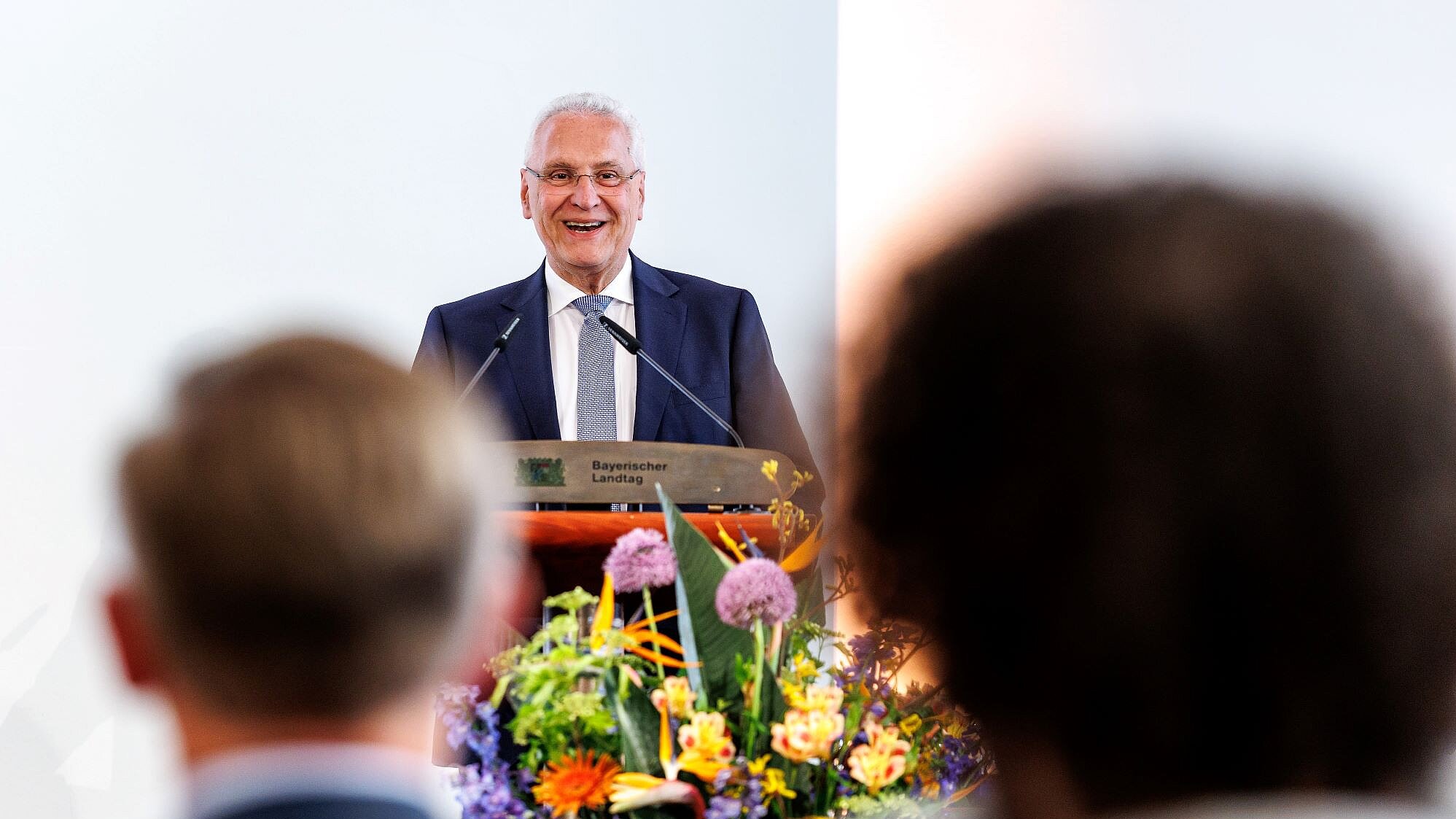 Innenminister Joachim Herrmann während seiner Rede am Rednerpult, vor ihm ein Blumendekor. Im Publikum sieht man Köpfe von hinten, die dem Minister zuhören.