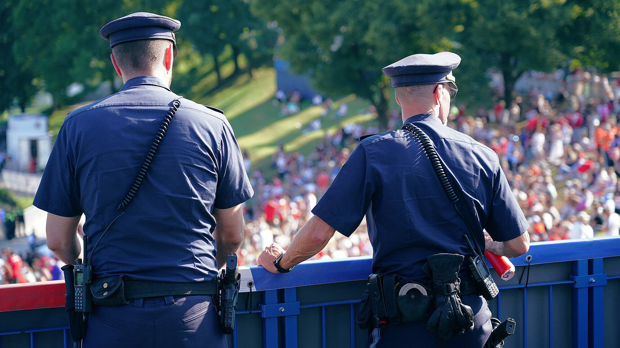 Zwei Polizeibeamte in Uniform stehen mit dem Rücken zur Kamera und beobachten eine im Freien versammelte Menschenmenge.